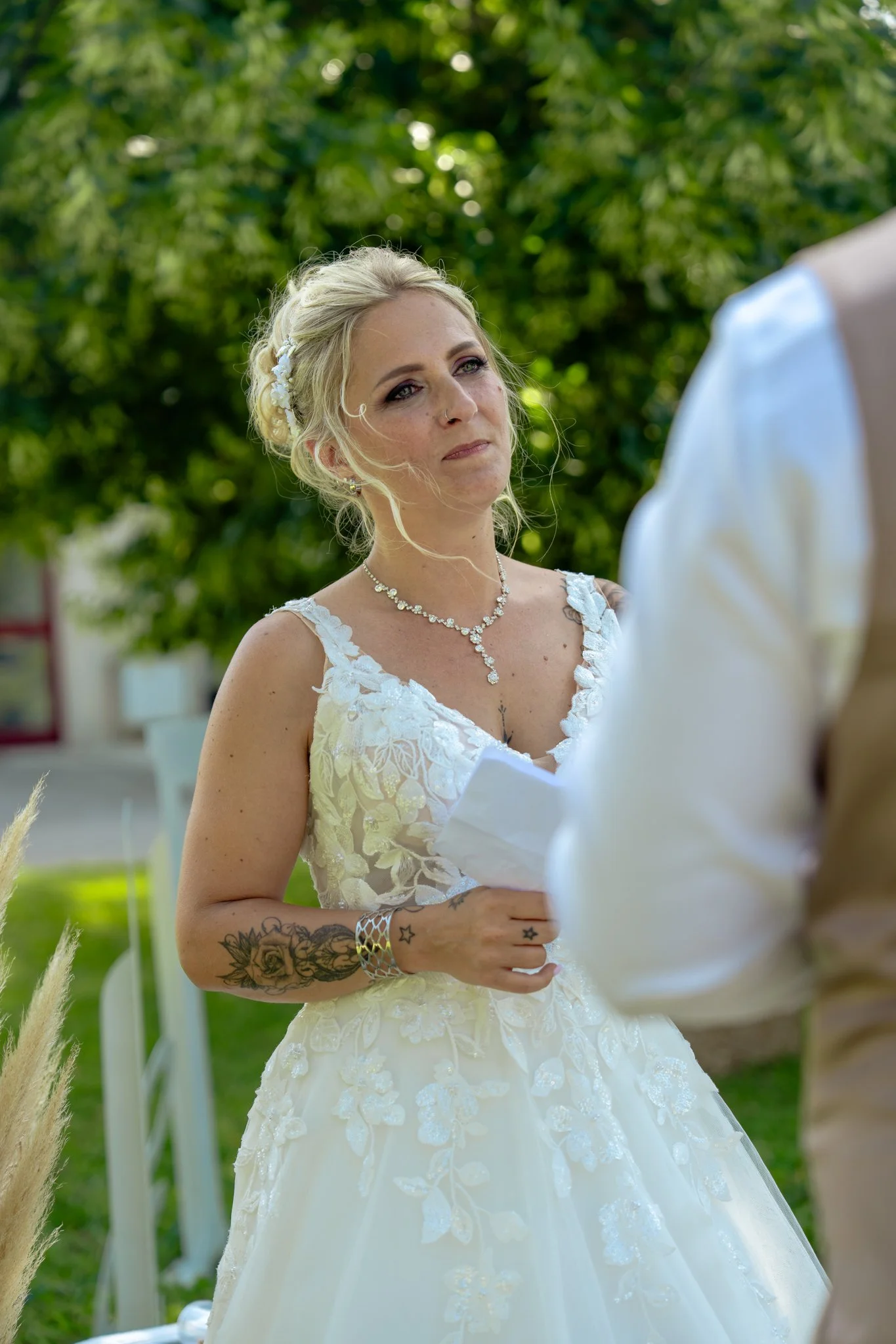 Une femme en robe de mariage lors d'une cérémonie en plein air, avec des cheveux blonds, portant un collier et une boucle d'oreilles.