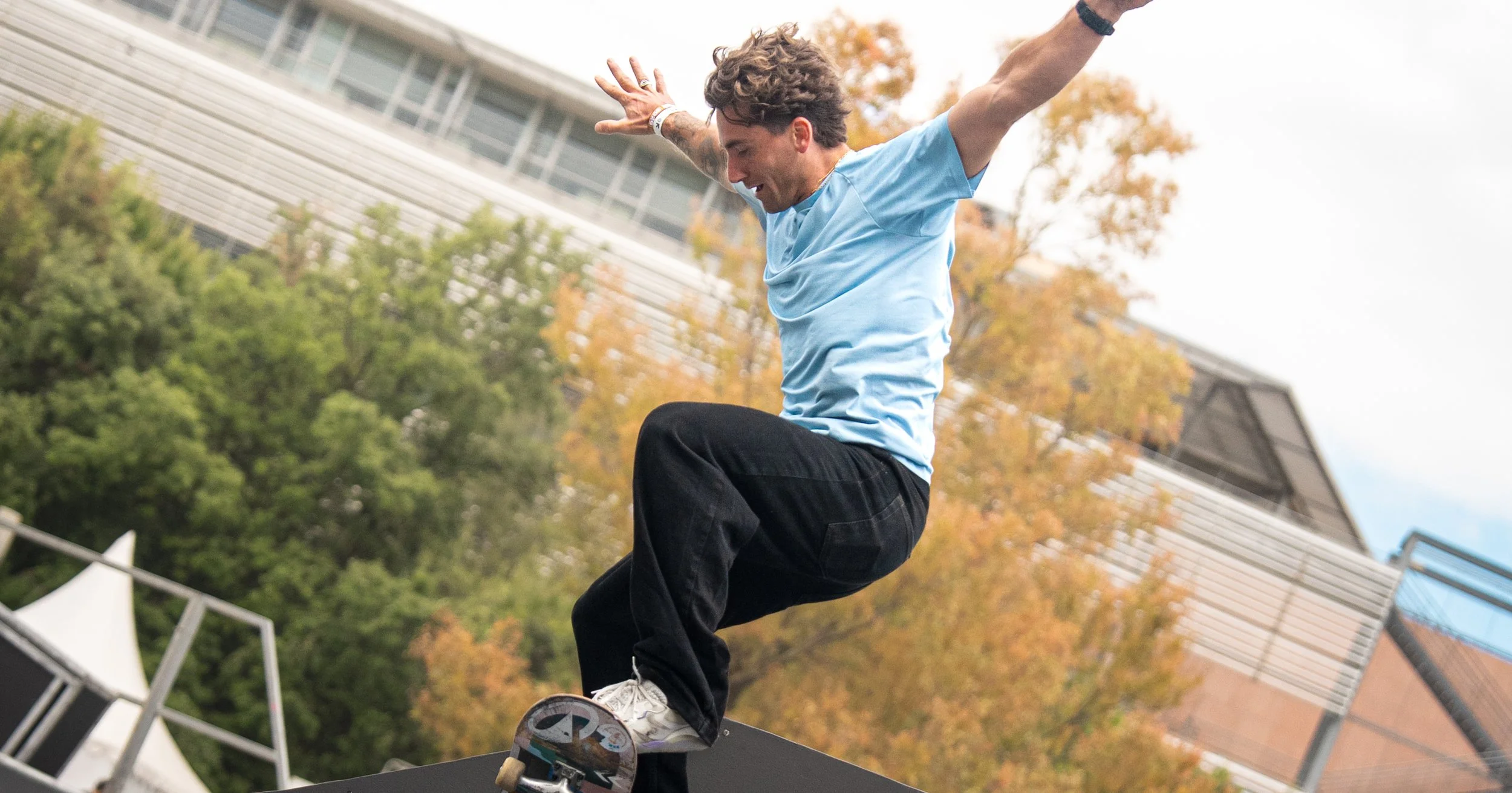 A young man skateboarding outdoors during autumn, wearing a light blue T-shirt and black pants, with trees changing colors and a modern building in the background.