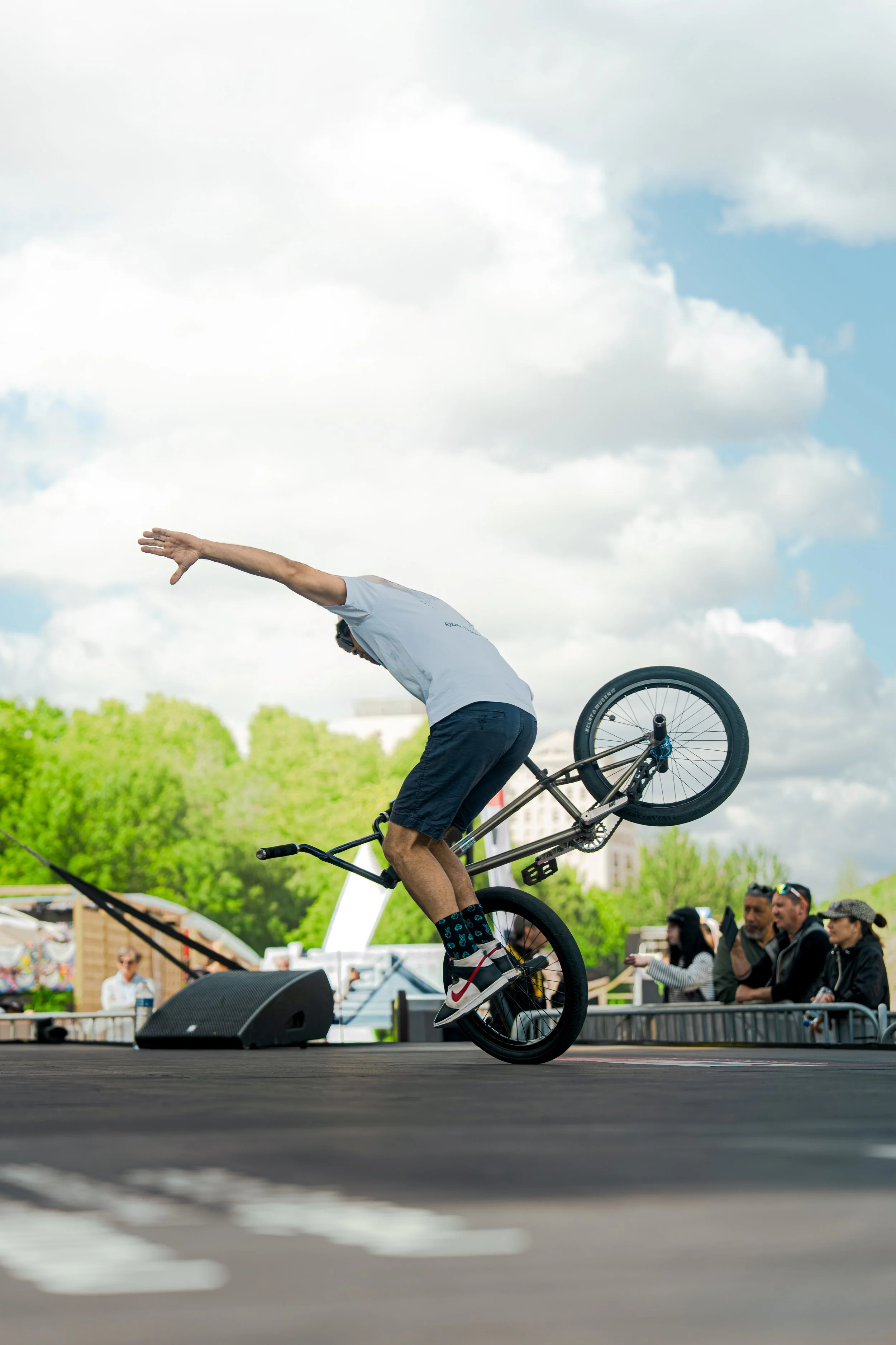 Un homme effectue une roue arrière sur un vélo lors d'un spectacle, avec un public en arrière-plan sous un ciel nuageux.