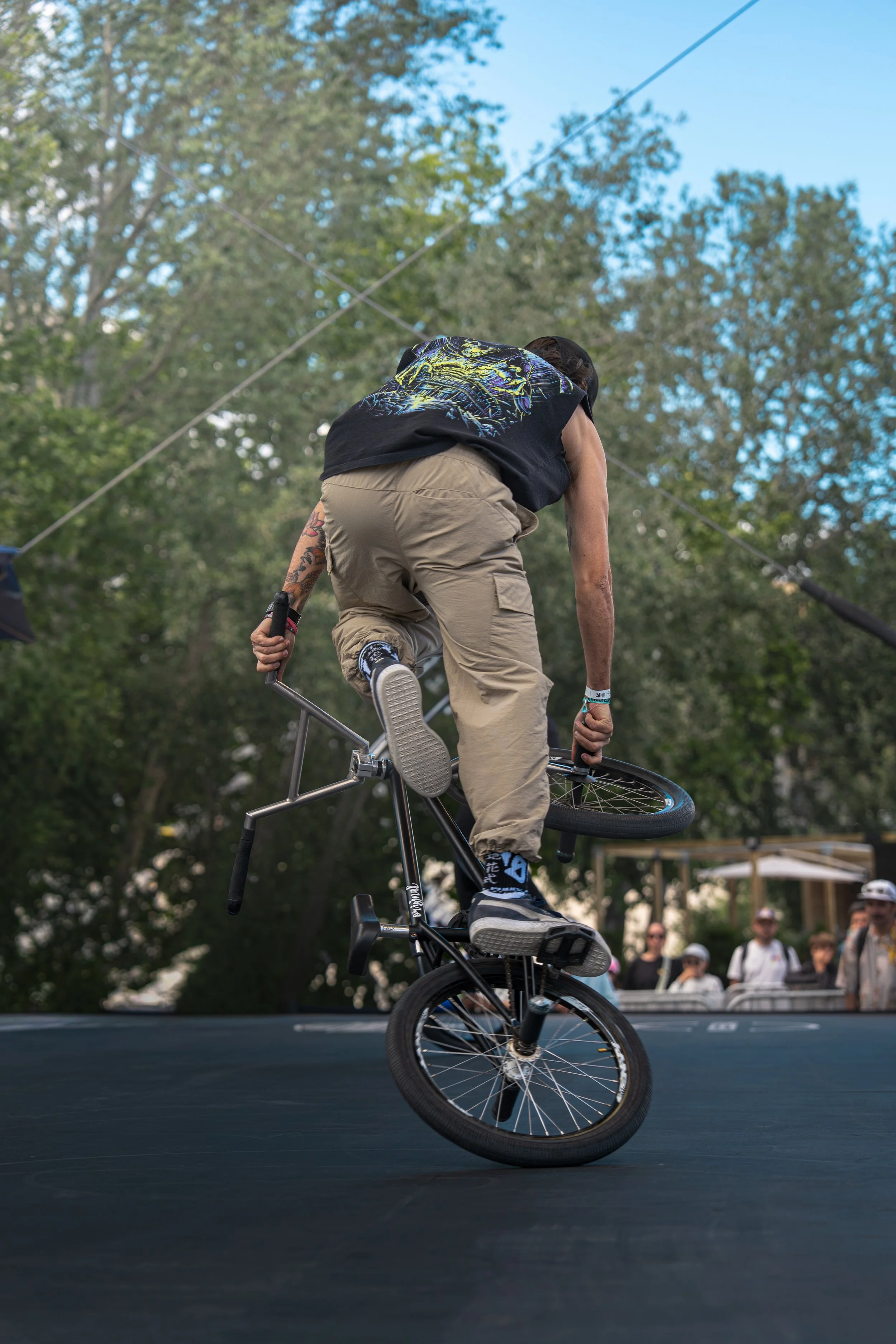 A person riding a BMX bike on a skate ramp during an outdoor event, with trees and spectators in the background.