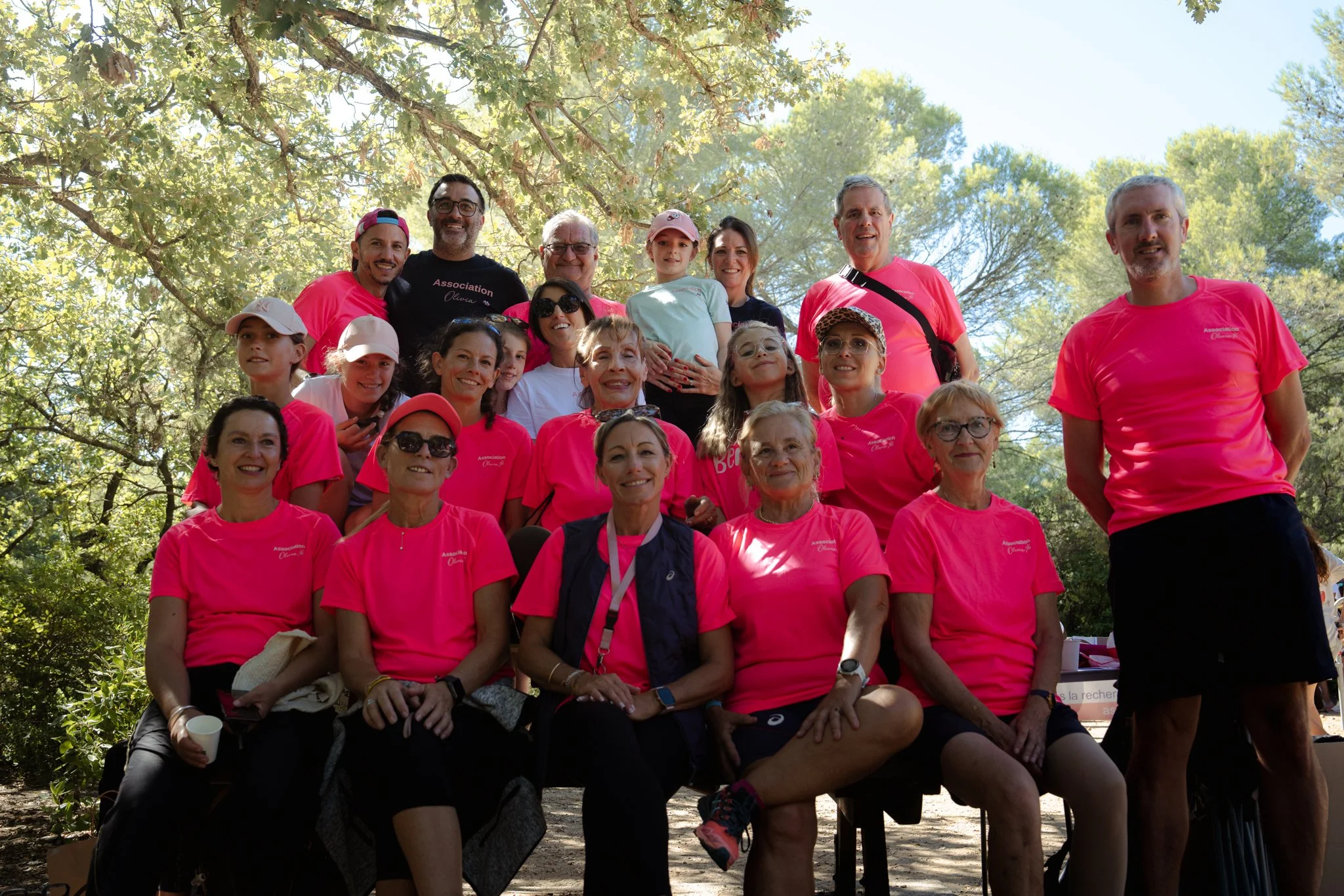 Groupe de personnes posant ensemble sous des arbres lors d'un événement en plein air. Beaucoup portent des t-shirts roses avec l'inscription 'Association'.