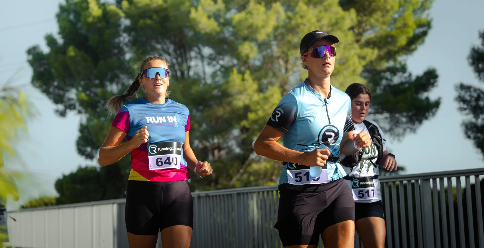 Trois femmes courent lors d'une course en plein air, portant des tenues de sport, des lunettes de soleil et des numéros de dossard. Arrière-plan de ciel bleu et de grands arbres verts.