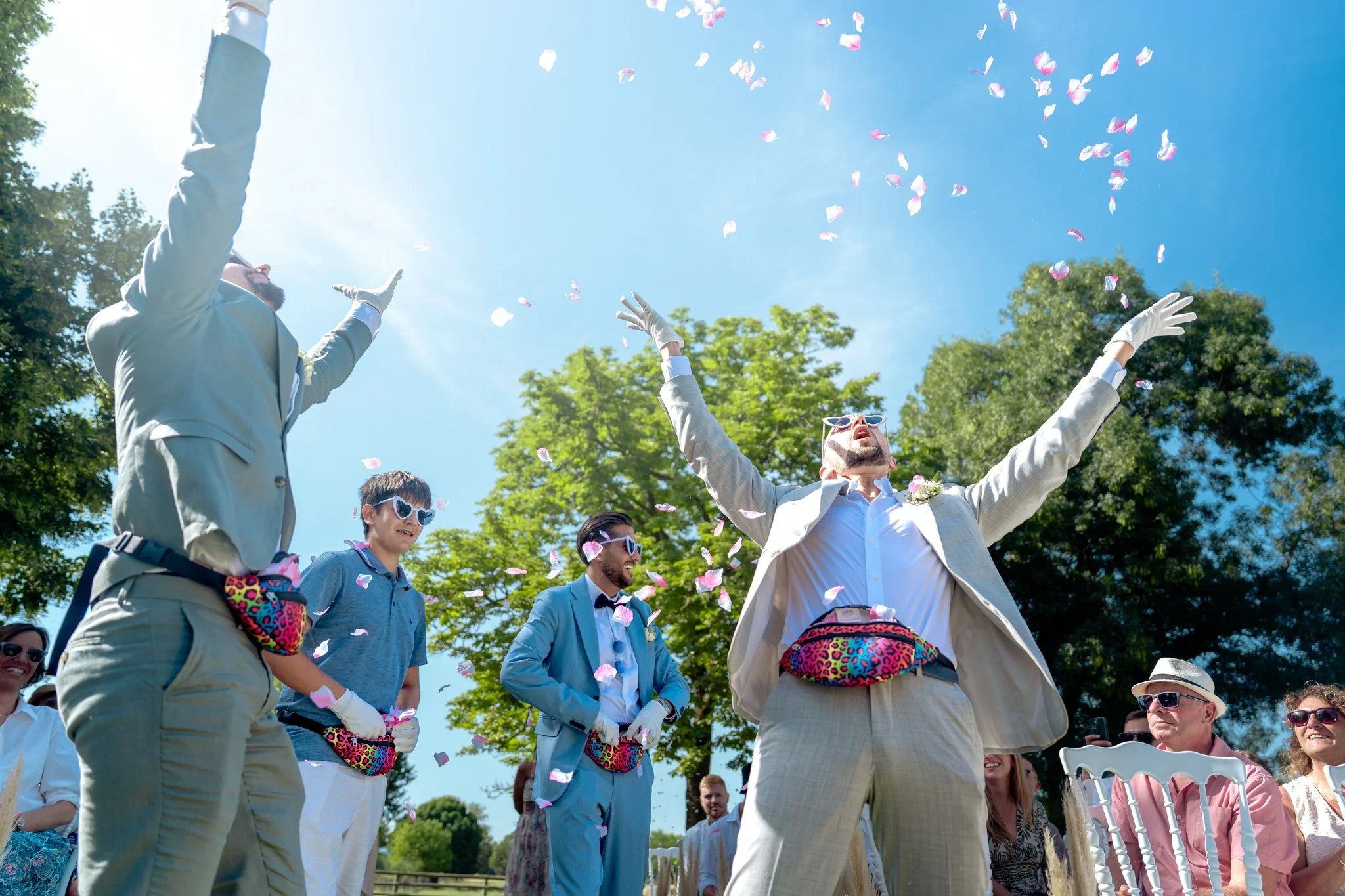 Un groupe de personnes lors d'un mariage en plein air, lançant des pétales de fleurs dans le ciel ensoleillé, avec des arbres en arrière-plan.