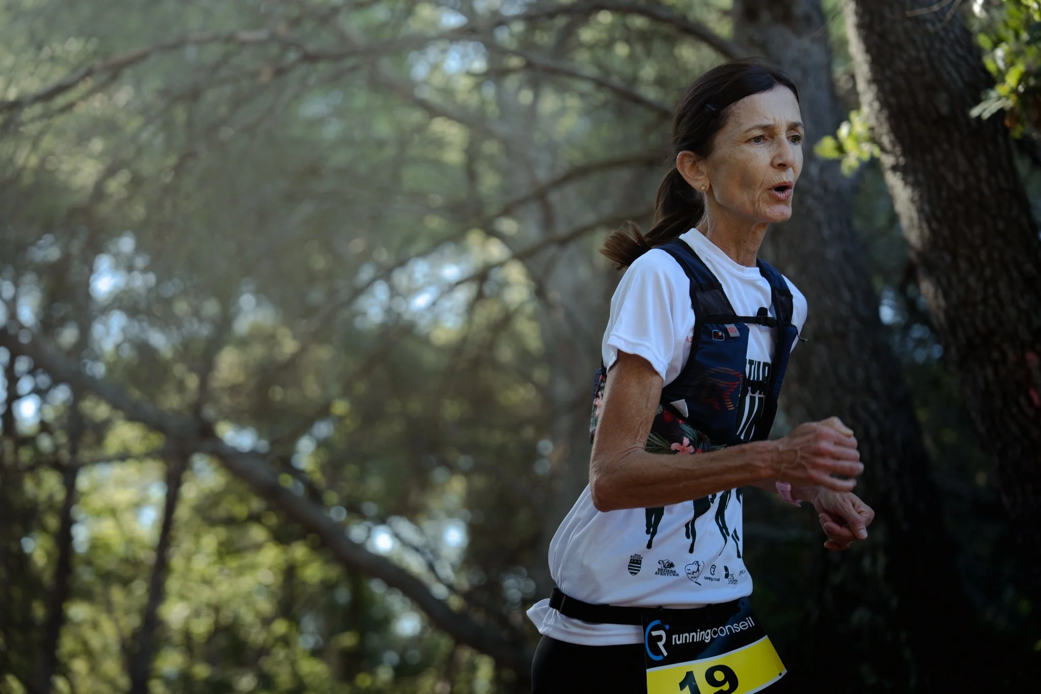 Une femme âgée court dans une forêt, portant un t-shirt blanc avec plusieurs logos, un sac à dos, et un dossard de course portant le numéro 19.