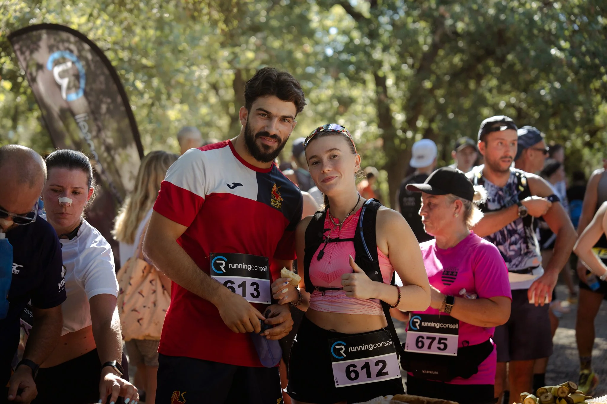 Participants de course marathon en plein air avec arbres et soleil en arrière-plan, portant des numéros de dossard, certains avec des boissons et équipements sportifs.