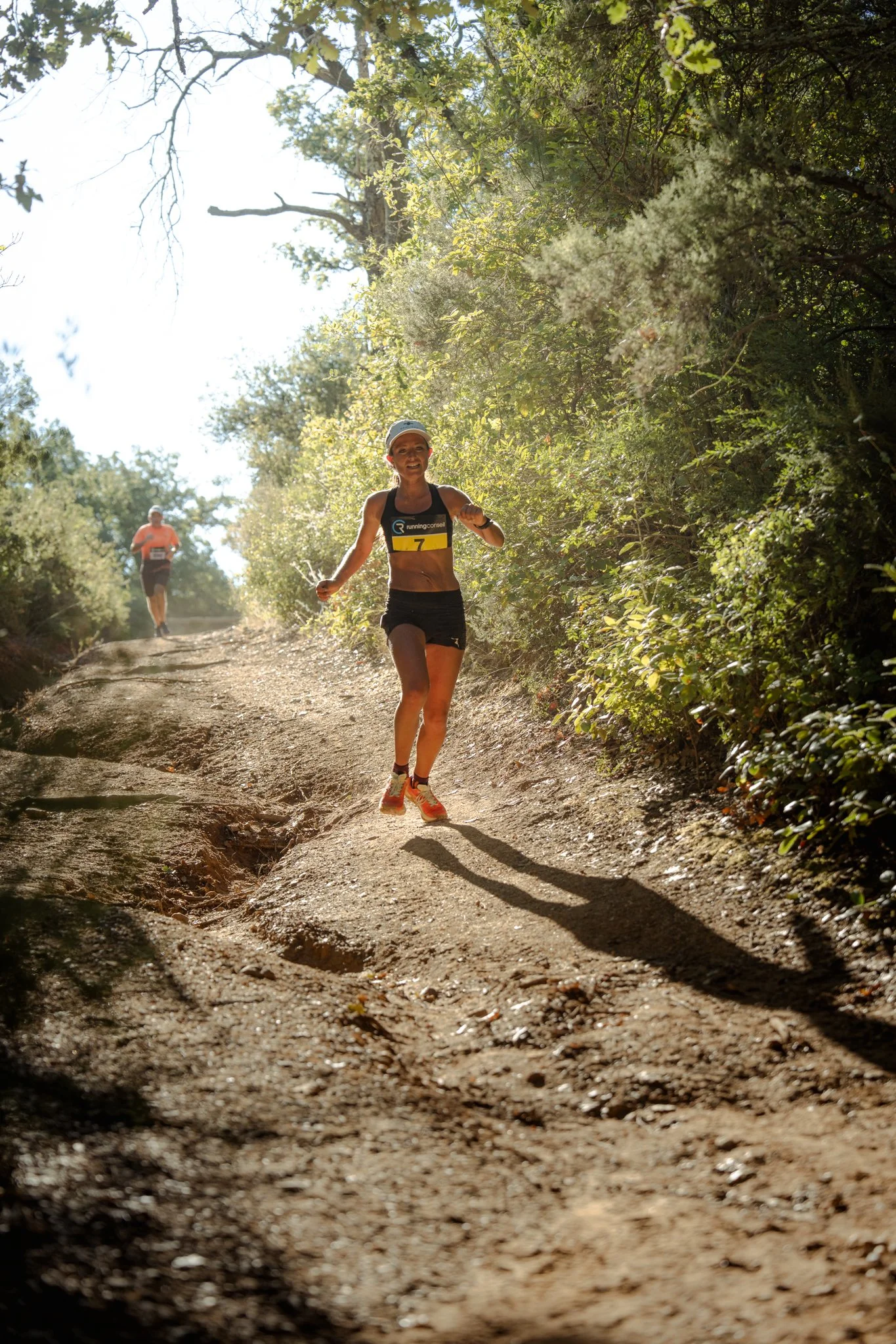 Deux coureurs, une femme et un homme, participant à une course en forêt, sur un sentier rocheux entouré de végétation dense, sous un ciel ensoleillé.