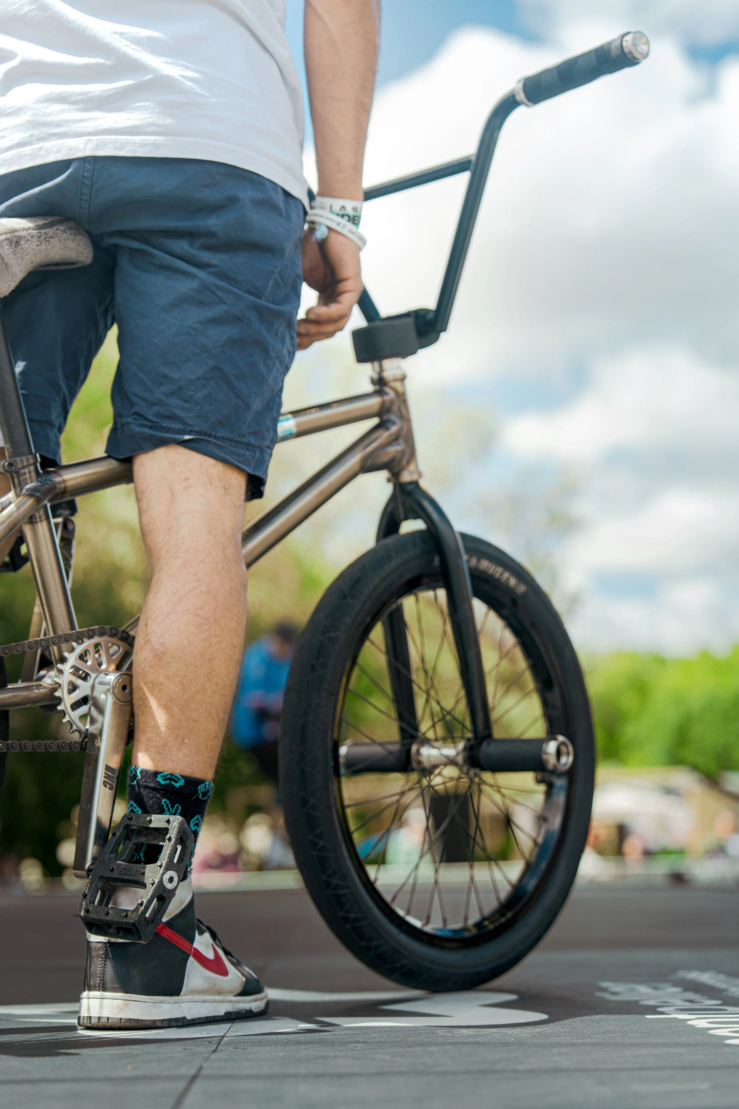 Un cycliste debout à côté de son vélo à l'extérieur, portant des chaussures de sport, une chemise blanche et un bracelet, avec un ciel partiellement nuageux en arrière-plan.
