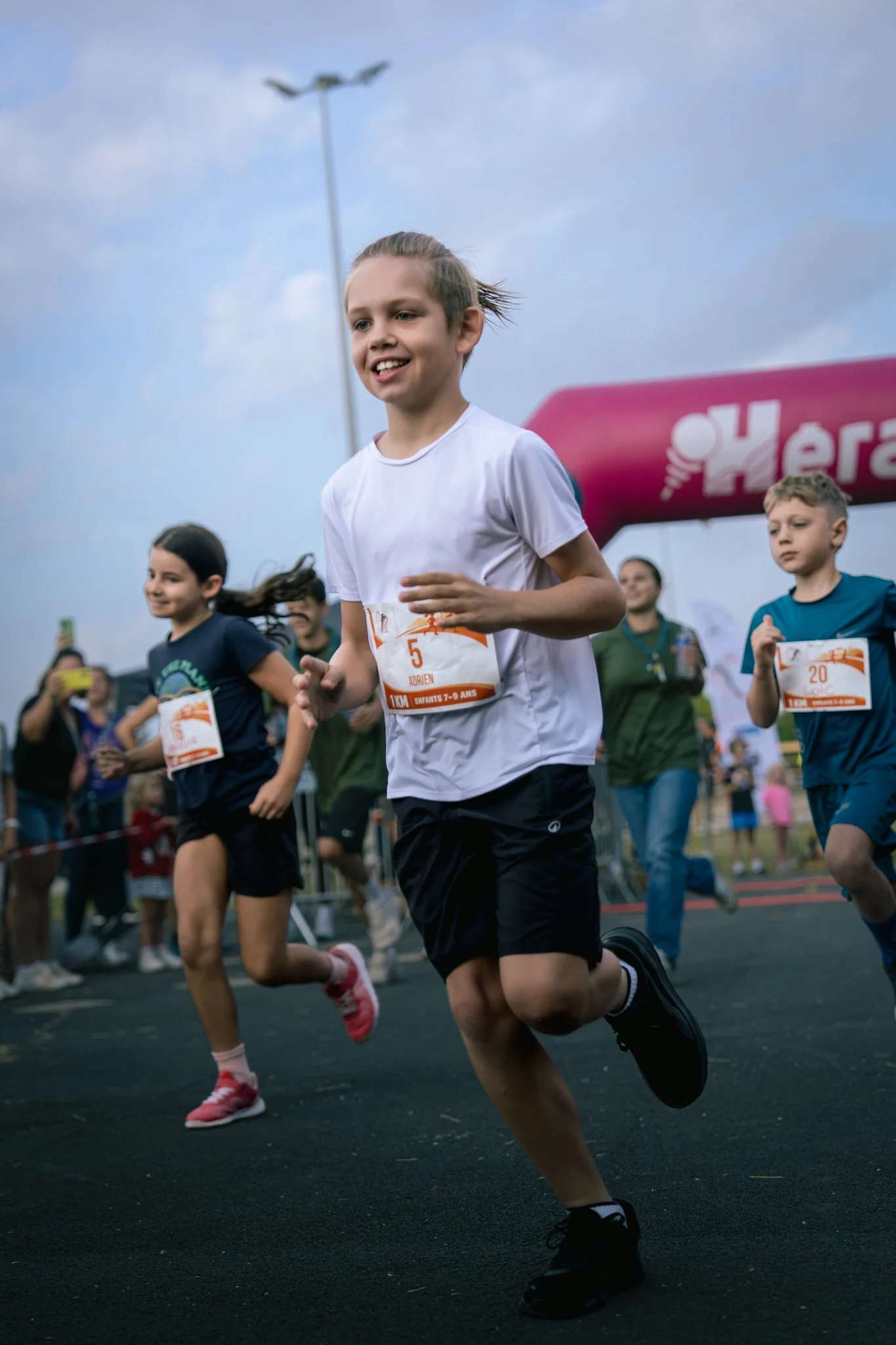 Des enfants courent lors d'une course, portant des numéros sur leurs vêtements, avec une foule de spectateurs en arrière-plan, sous un ciel nuageux.