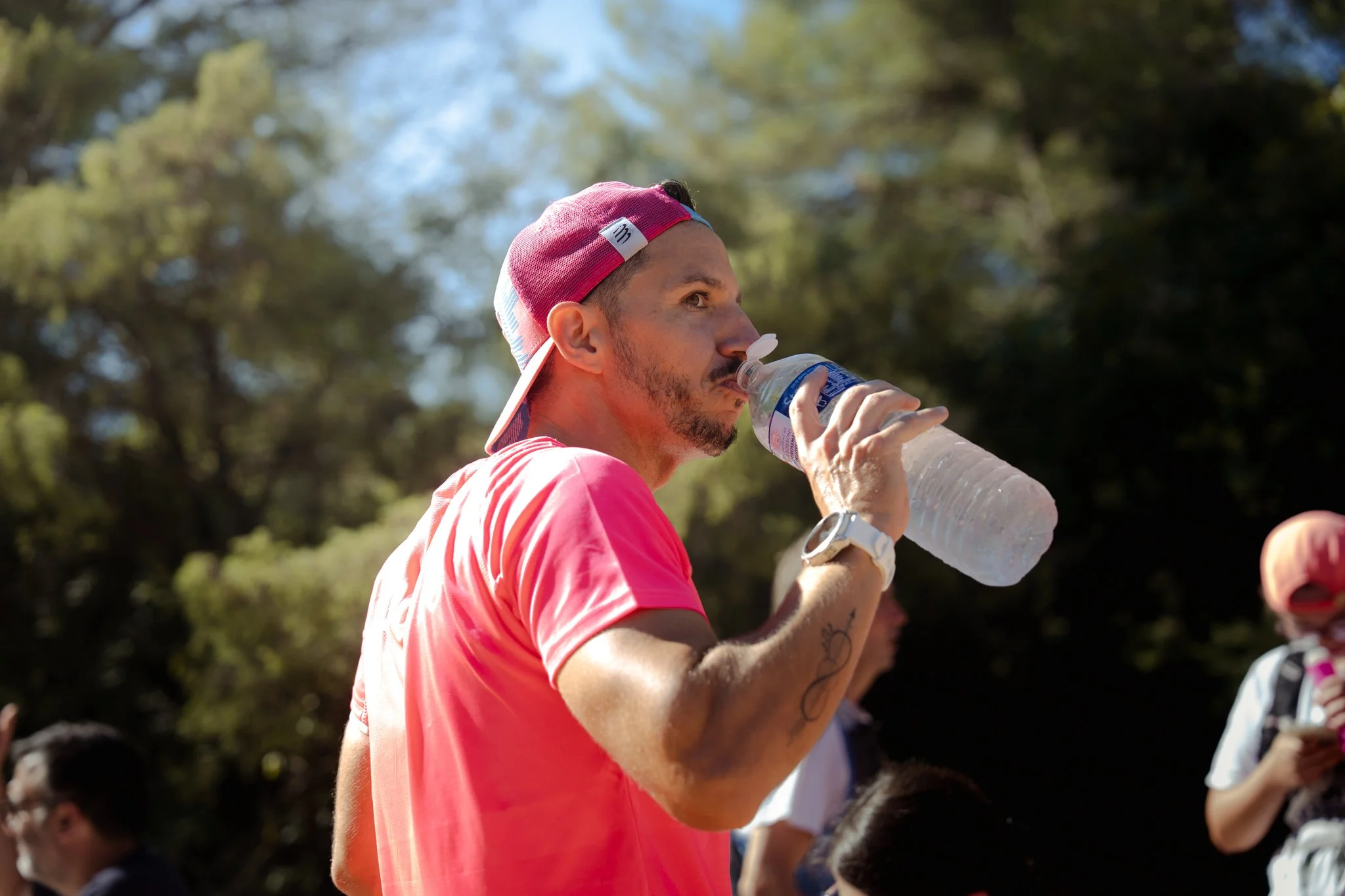 Homme portant un t-shirt rose et un bonnet rose buvant de l'eau lors d'une activité en plein air. Arrière-plan de feuillage et de ciel bleu.