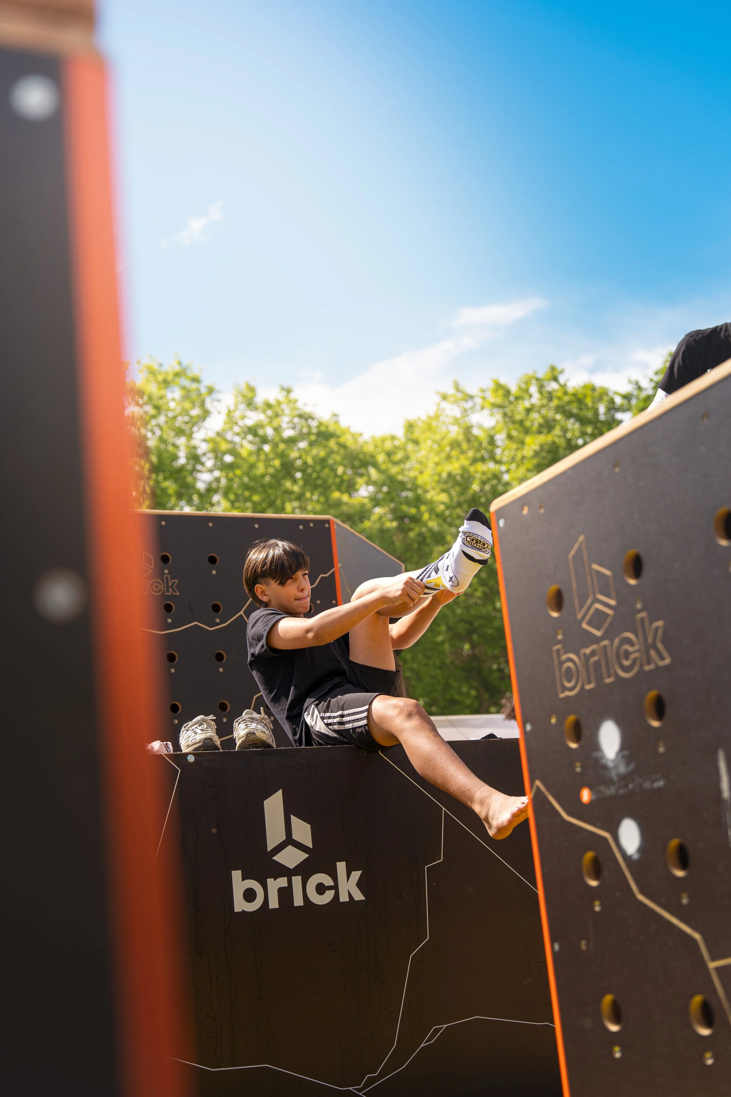 Un jeune garçon escalade une structure d'escalade en plein air par une journée ensoleillée, avec un ciel bleu clair et des arbres verts en arrière-plan.