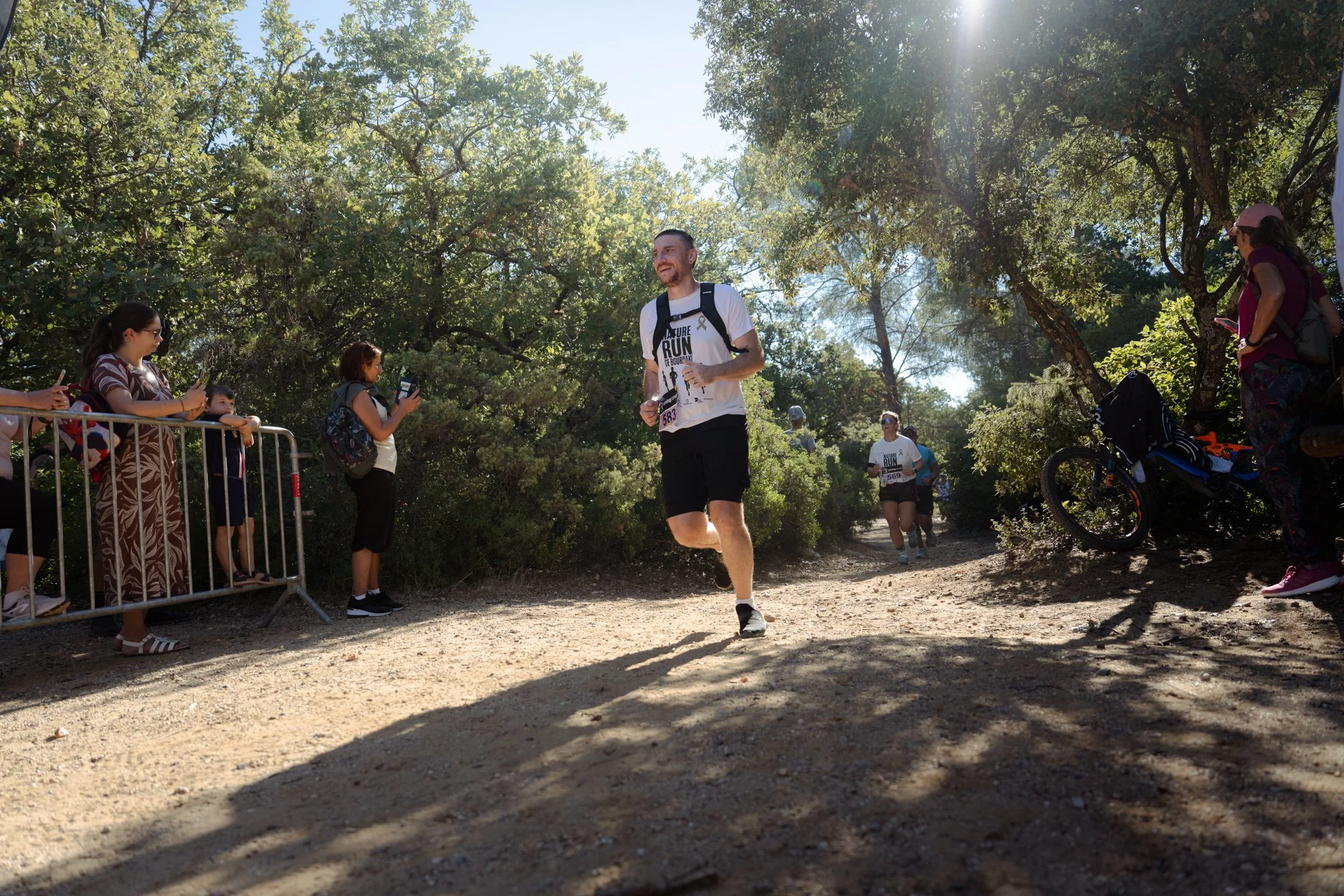 Un homme participant à une course ou un trail dans une forêt, entouré de spectateurs et de cyclistes, sous un ciel ensoleillé.