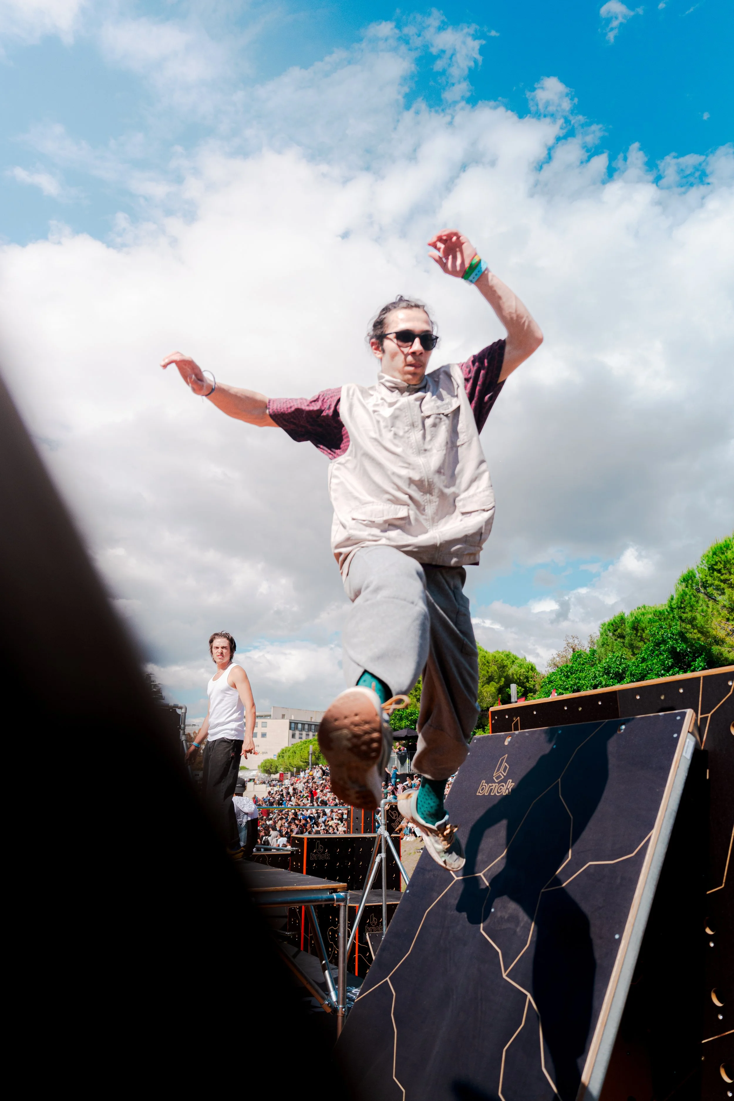 Un homme portant des lunettes de soleil, un t-shirt à manches courtes et un gilet beige saute lors d'un événement en plein air avec un public nombreux et un ciel partiellement nuageux.