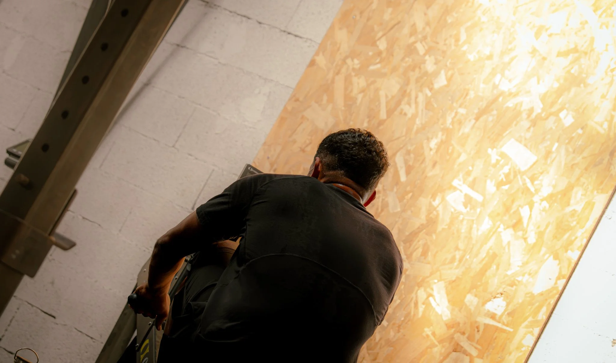 Un homme vu de dos, assis ou accroupi, dans une pièce avec un mur en panneau de particules de bois et un mur en blocs de béton. Il semble occupé à une activité, peut-être à utiliser un ordinateur portable.