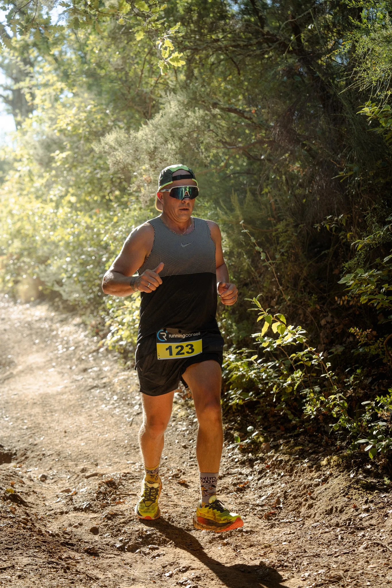 Un homme court sur un sentier en forêt, portant un t-shirt de sport gris et noir, un chapeau, des lunettes de navigation, un maillot de course numéro 123, des chaussettes colorées et des chaussures de course jaunes. Le soleil filtre à travers les arb