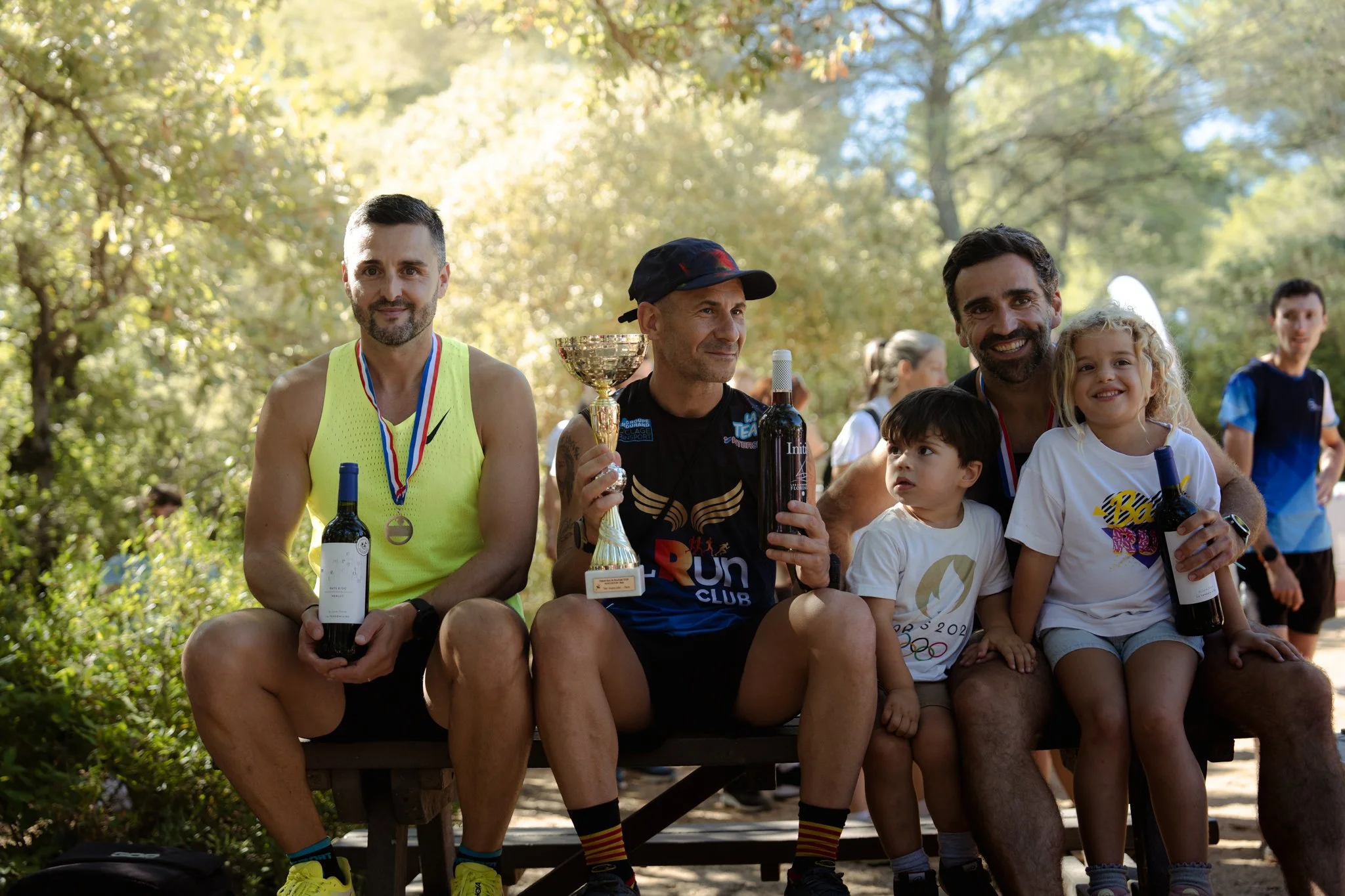 Un groupe d'hommes et d'enfants posant avec des médailles, des coupes et des bouteilles de vin après une course ou un événement sportif dans un parc en automne.