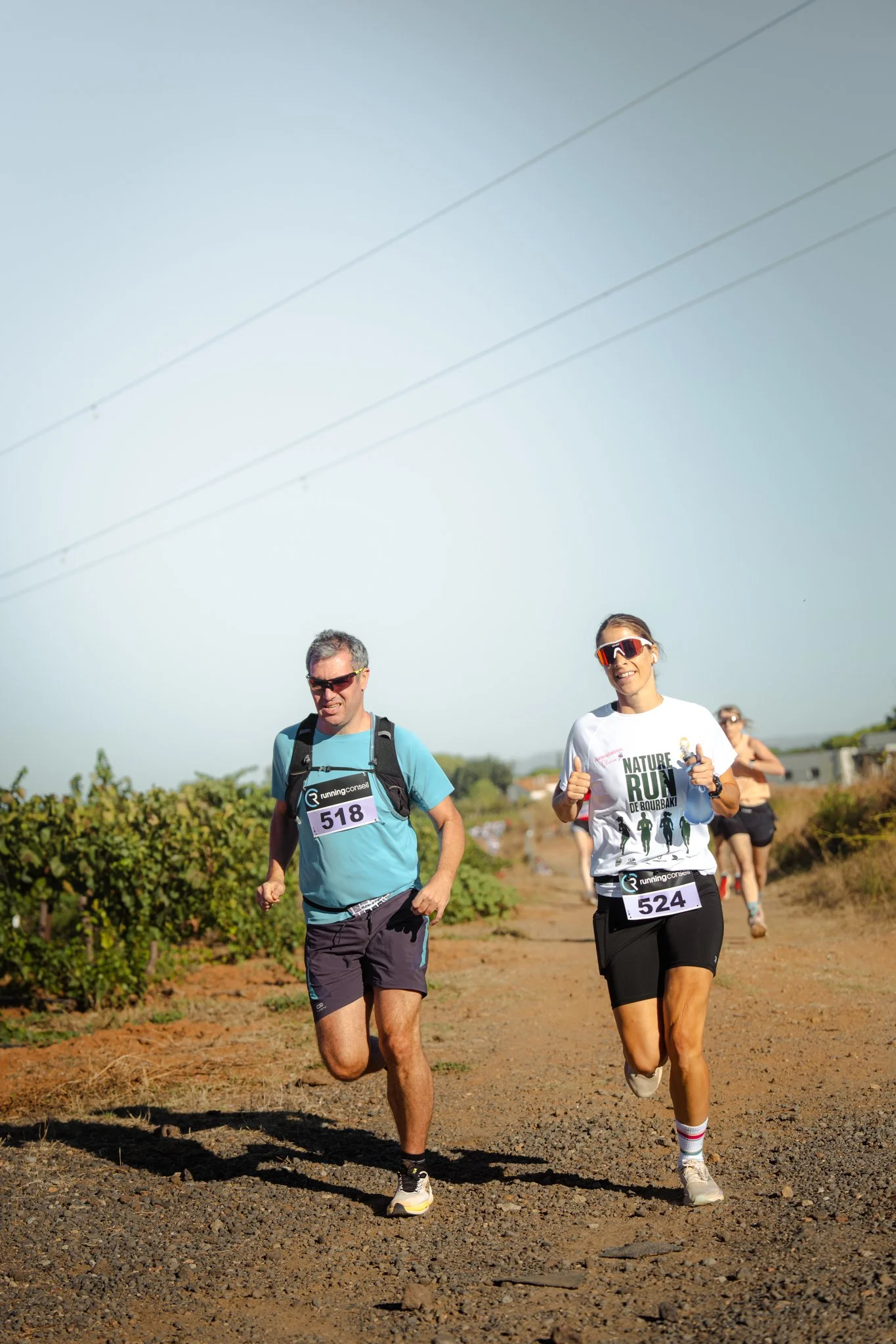 Deux coureurs participant à une course, portant des dossards 518 et 524, courent sur un chemin en extérieur, avec un arrière-plan comprenant des buissons et un ciel clair.