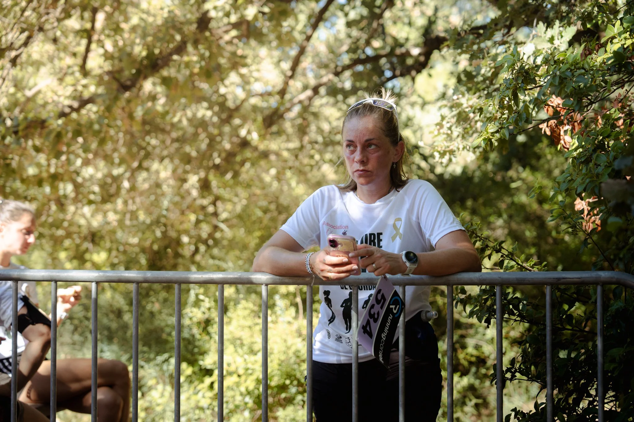 Une femme regarde son téléphone alors qu'elle est assise derrière une barrière en extérieur, avec des arbres en arrière-plan, un homme et une autre femme sont également présents.
