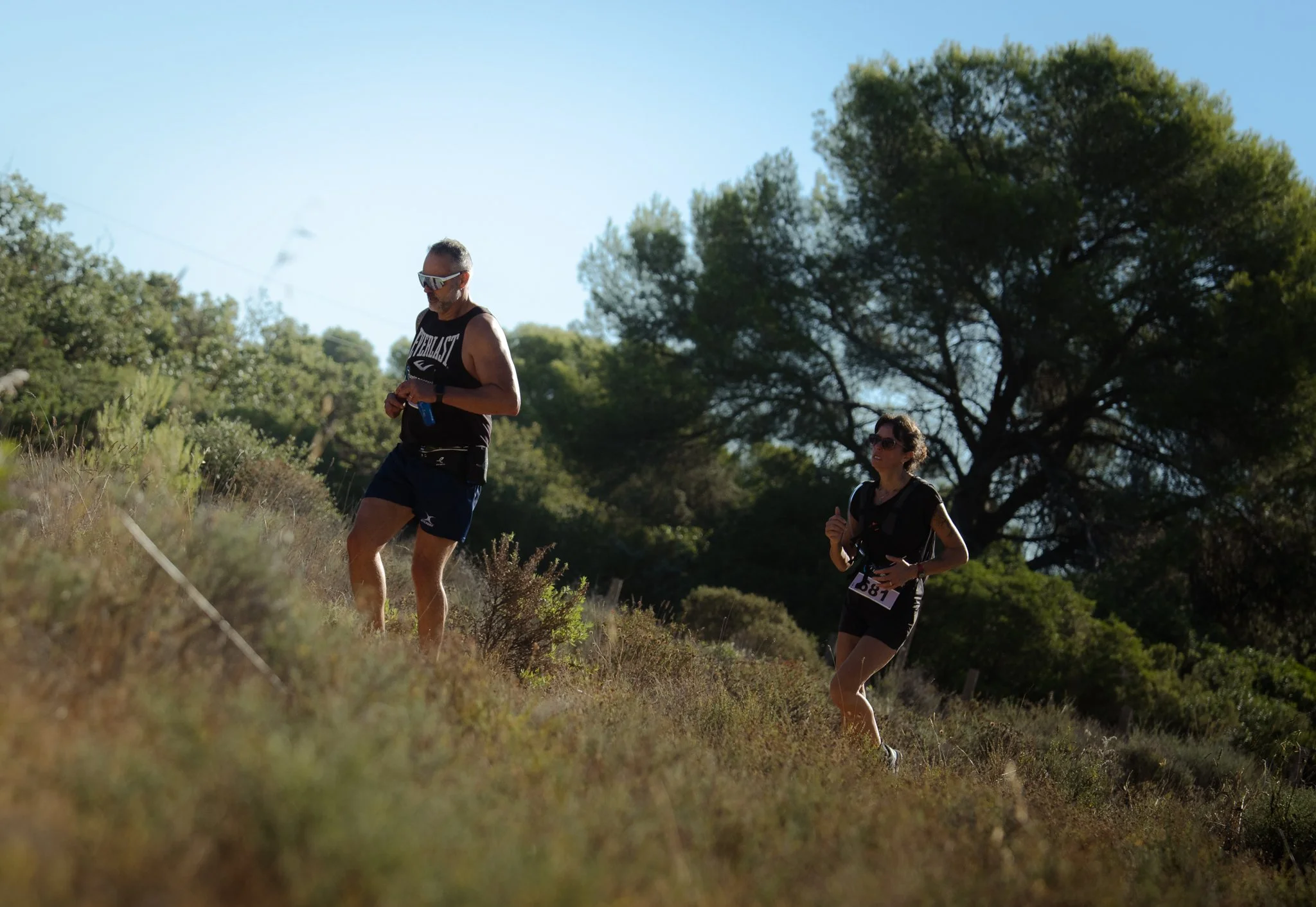 Deux personnes faisant de la course à pied ou de la marche dans un espace naturel avec des arbres, sous un ciel clair et ensoleillé.