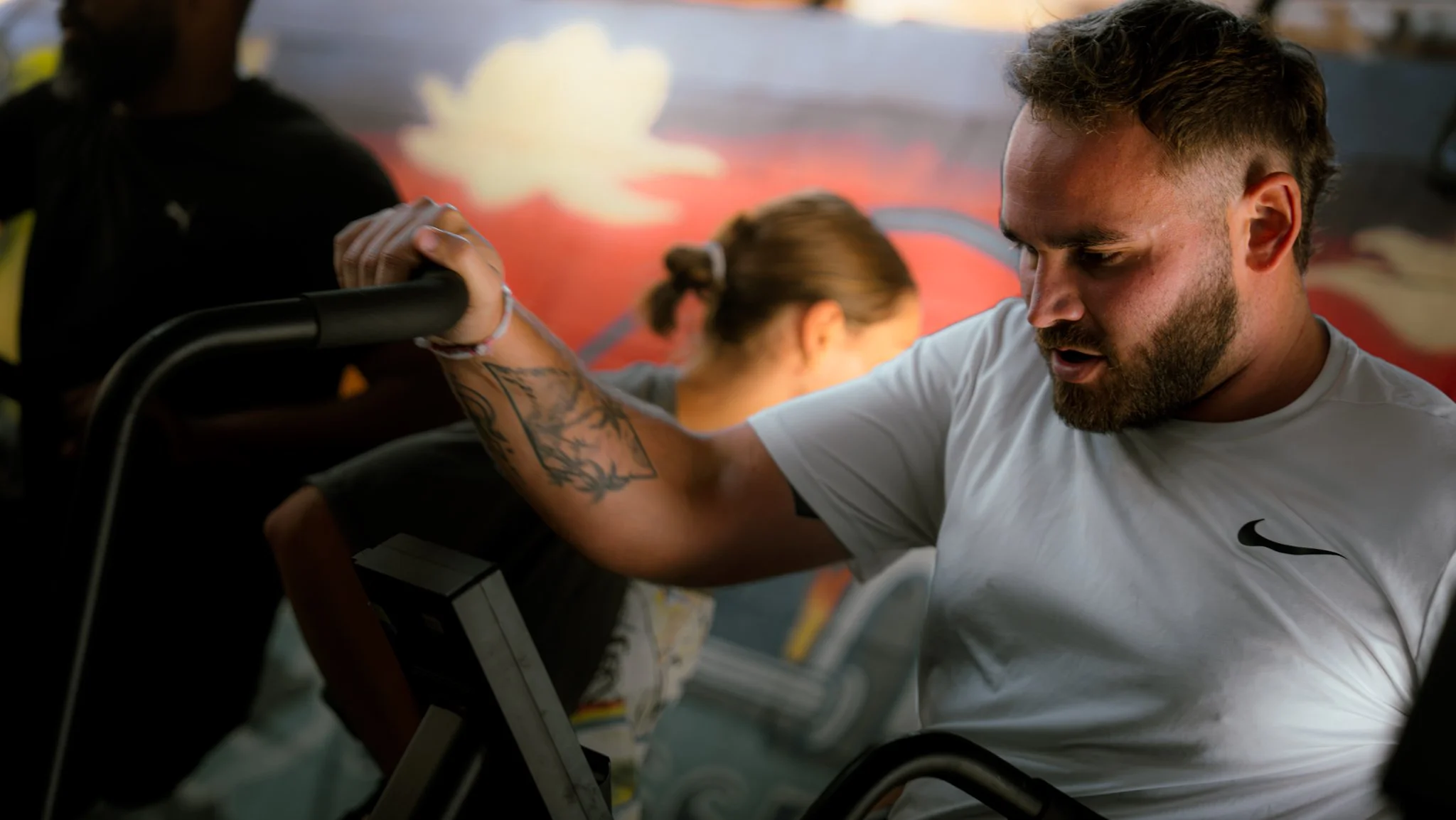 Un homme fait un exercice de musculation en utilisant une machine à haltères, dans une salle de sport, avec un fond en dégradé de couleurs chaudes et une silhouette floue