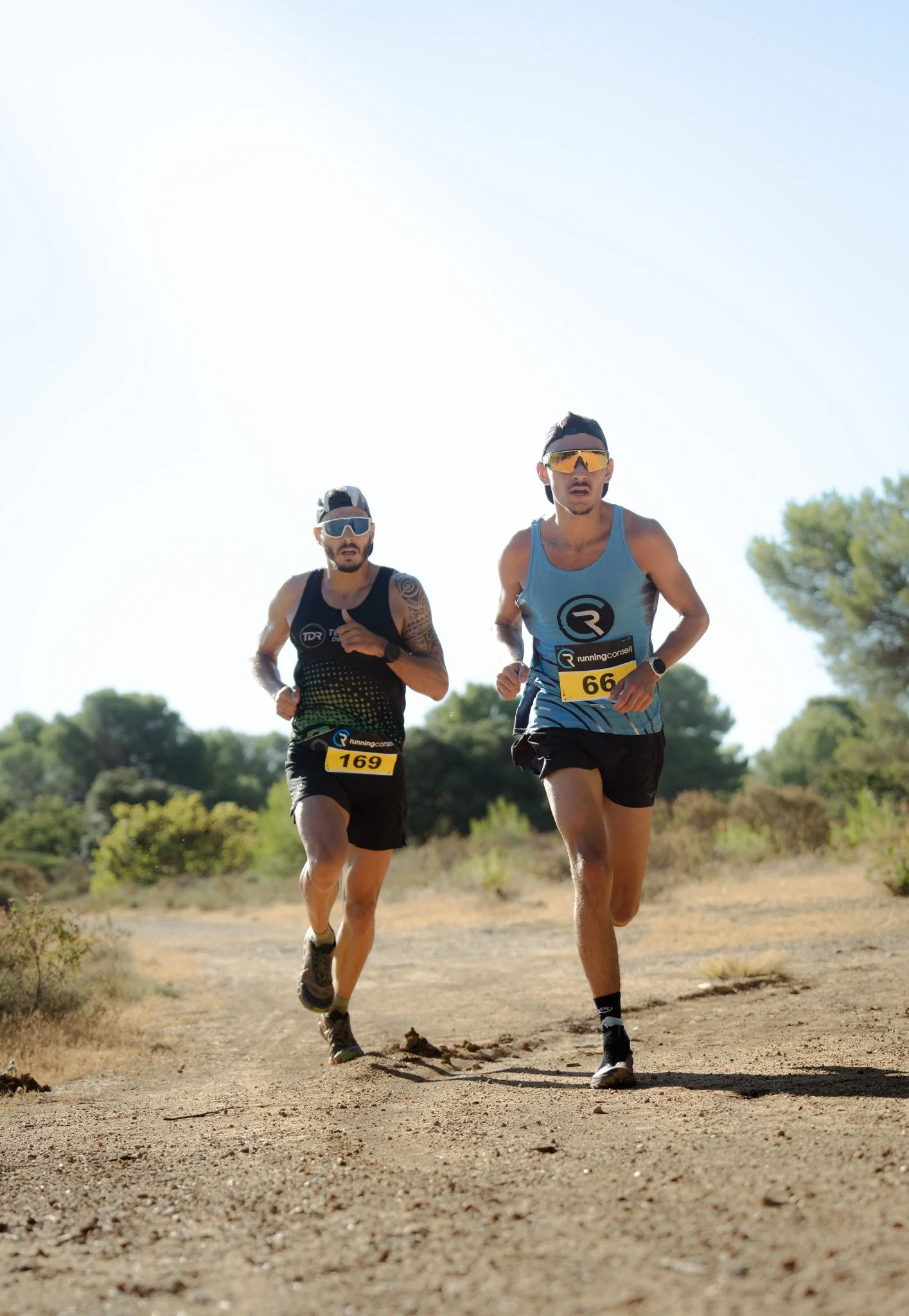 Deux hommes courent sur un sentier en plein air, portant des vêtements de course, des lunettes de soleil, des casquettes et des chiffres de course, avec un fond de végétation et un ciel clair.