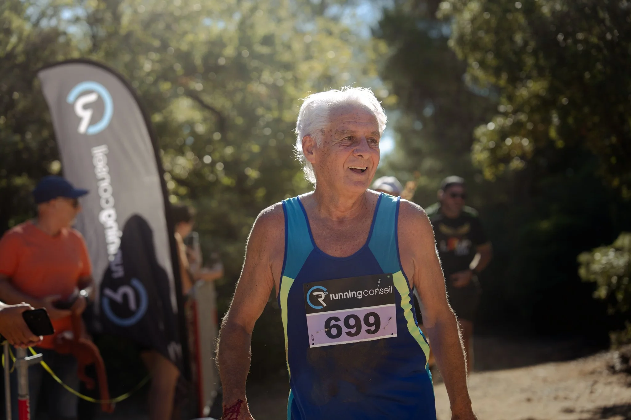 Un homme âgé participe à une course en plein air, portant un maillot bleu avec une dossard numéro 599, entouré d'autres coureurs et spectateurs sous un ciel ensoleillé.