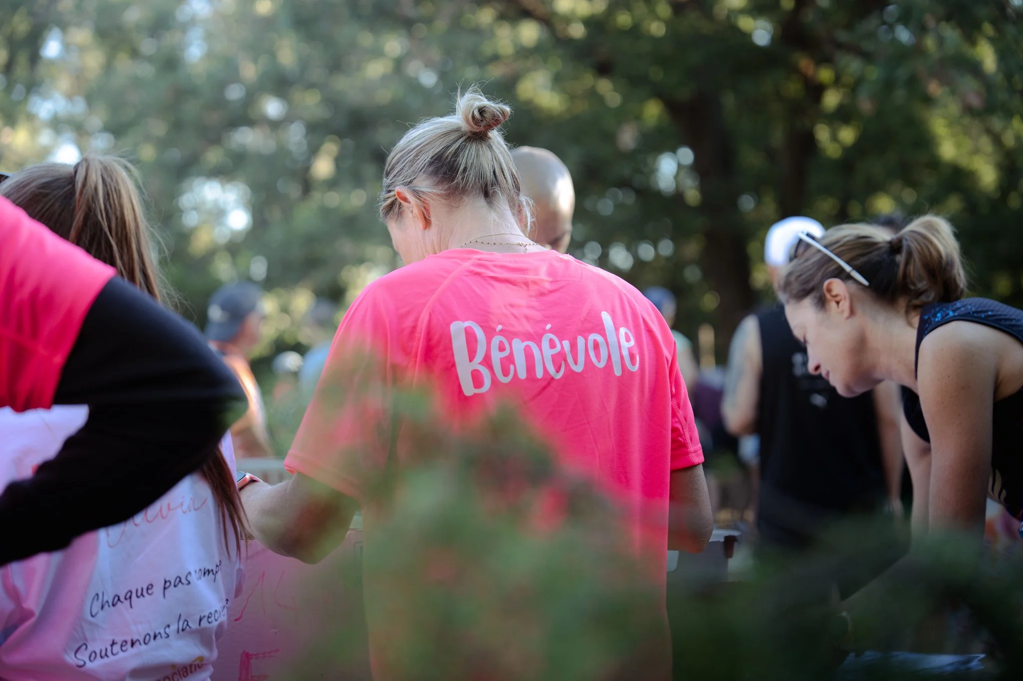 Groupe de personnes, principalement des femmes, participent à un événement en plein air, portant des vêtements roses et avec des textes imprimés, dans un environnement boisé. La personne au centre porte un T-shirt rose avec l'inscription "Bénévole" s