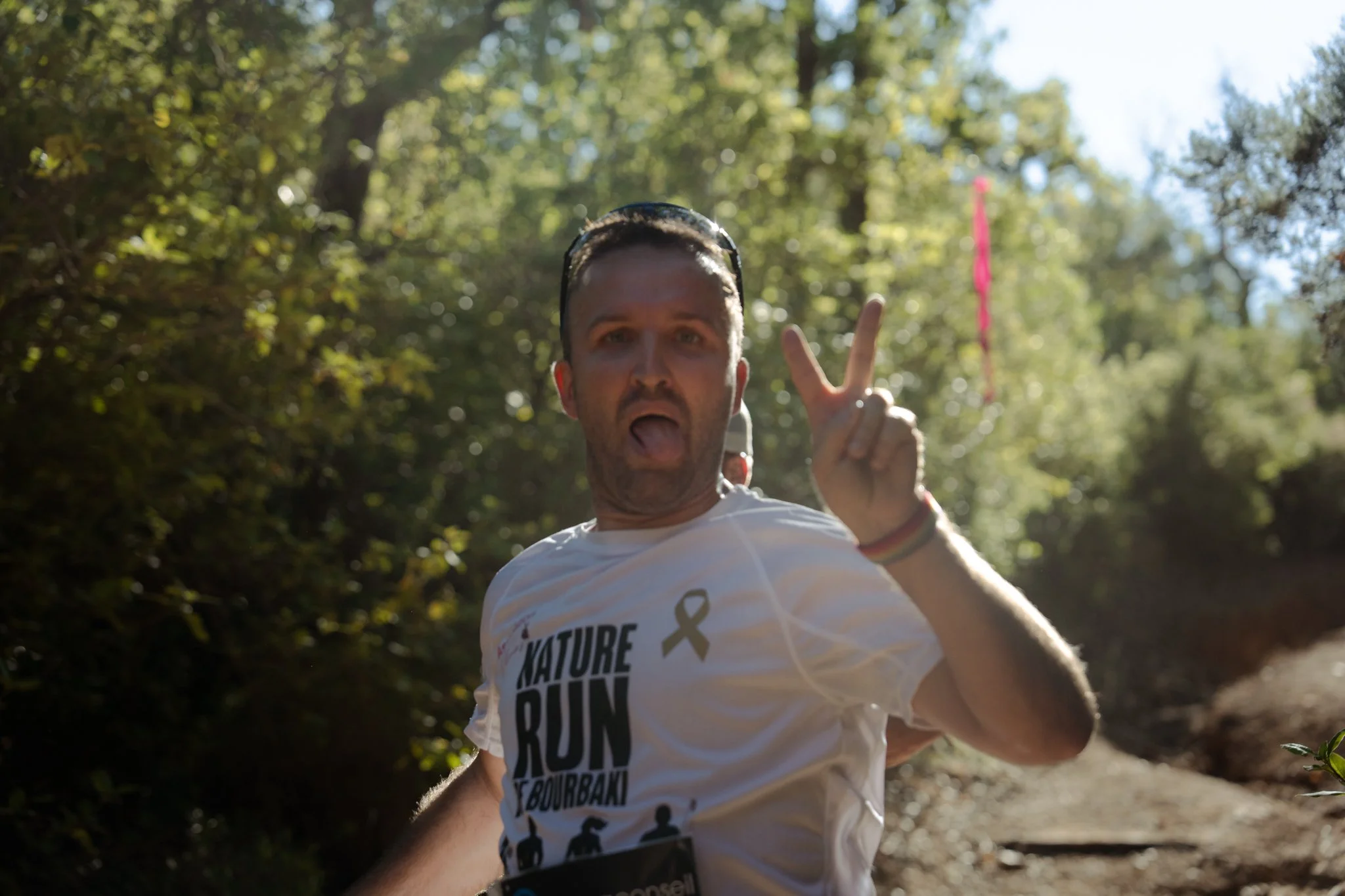 Homme faisant un geste de paix avec deux doigts lors d'une course dans un parc ensoleillé, portant un t-shirt blanc avec des écritures et des silhouettes de coureurs.