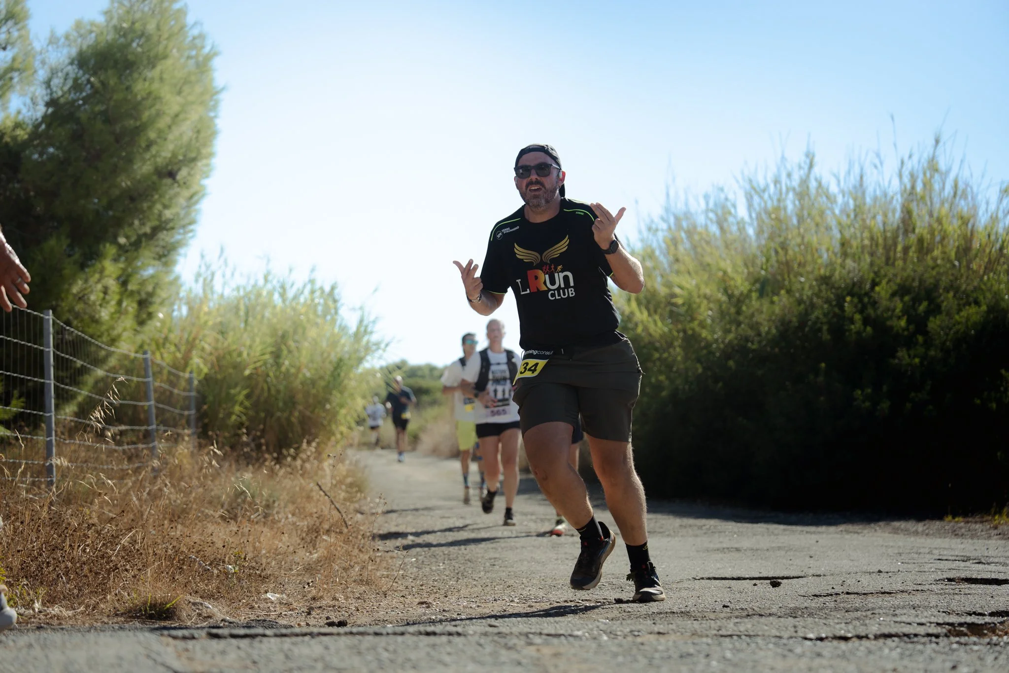 Un homme qui court, portant un t-shirt noir avec un logo de course, des lunettes de soleil, et une casquette, participe à une course sur un sentier en plein air avec d'autres coureurs en arrière-plan.