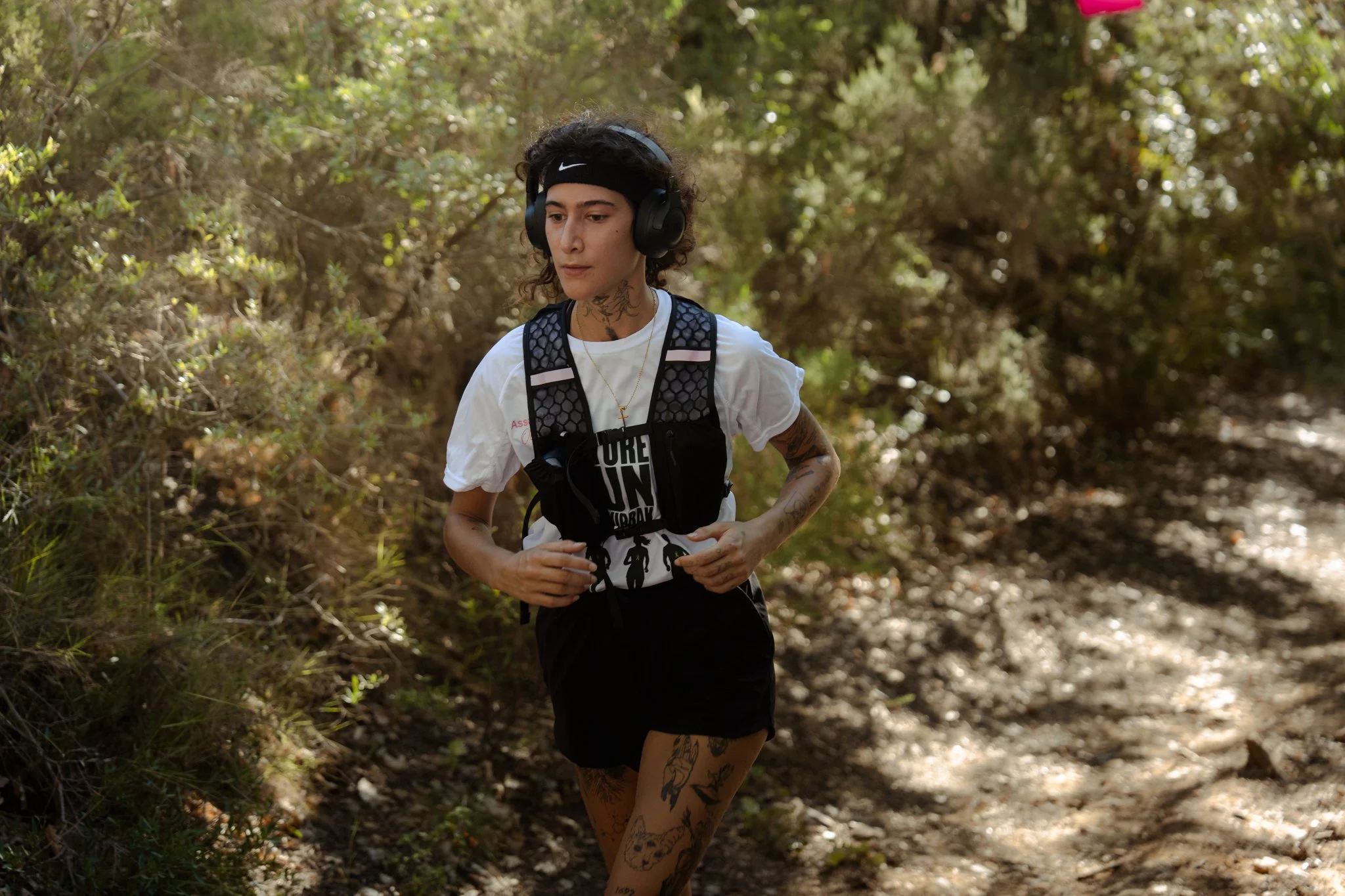 Une jeune femme avec des tatouages court vêtue court, portant un sac à dos et une paire d'écouteurs, court sur un sentier forestier.