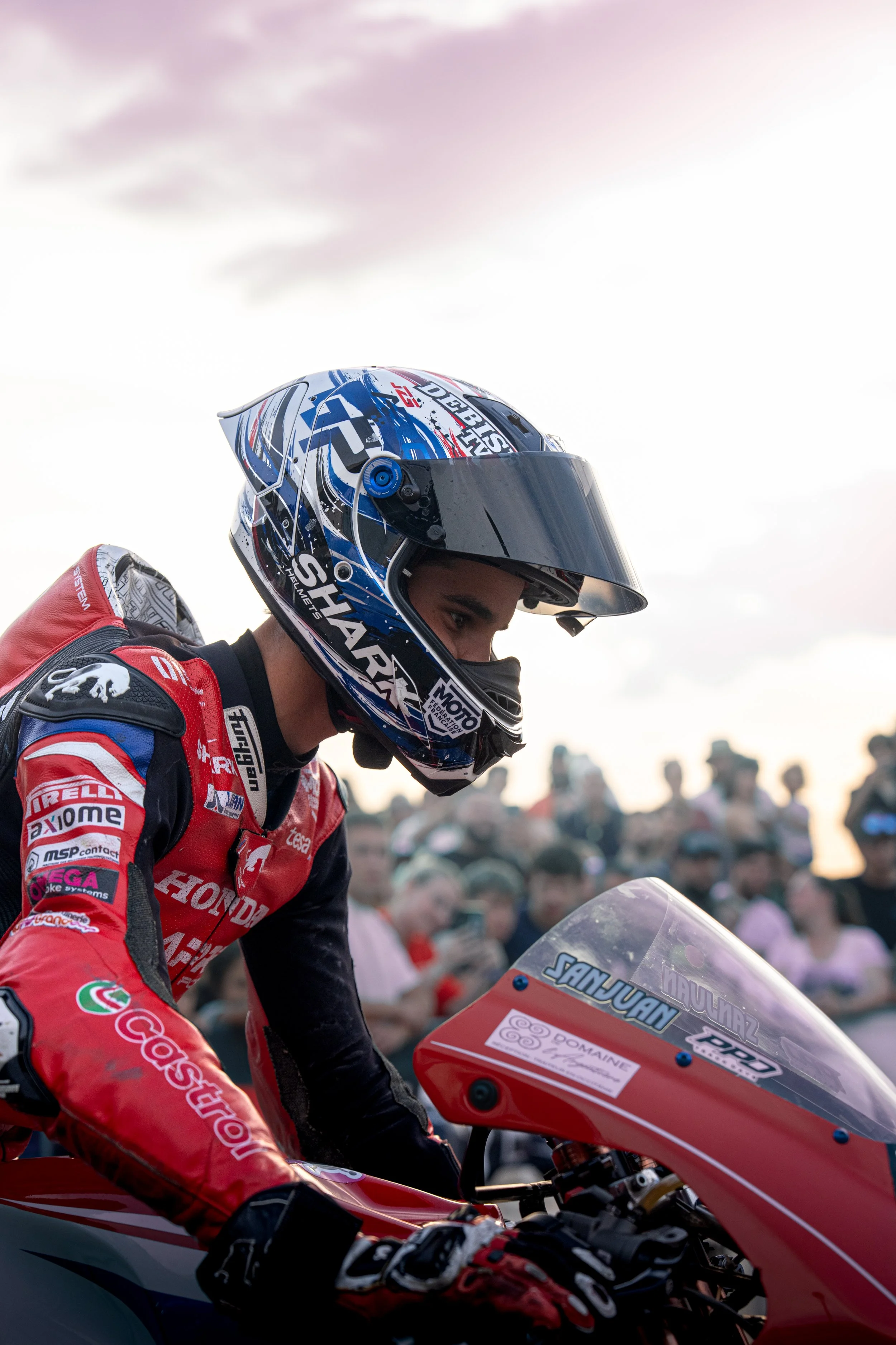 A motorcycle racer in a red racing suit and a blue, white, and black helmet is sitting on a red motorcycle. The background shows a crowd of spectators watching outdoors at dusk.