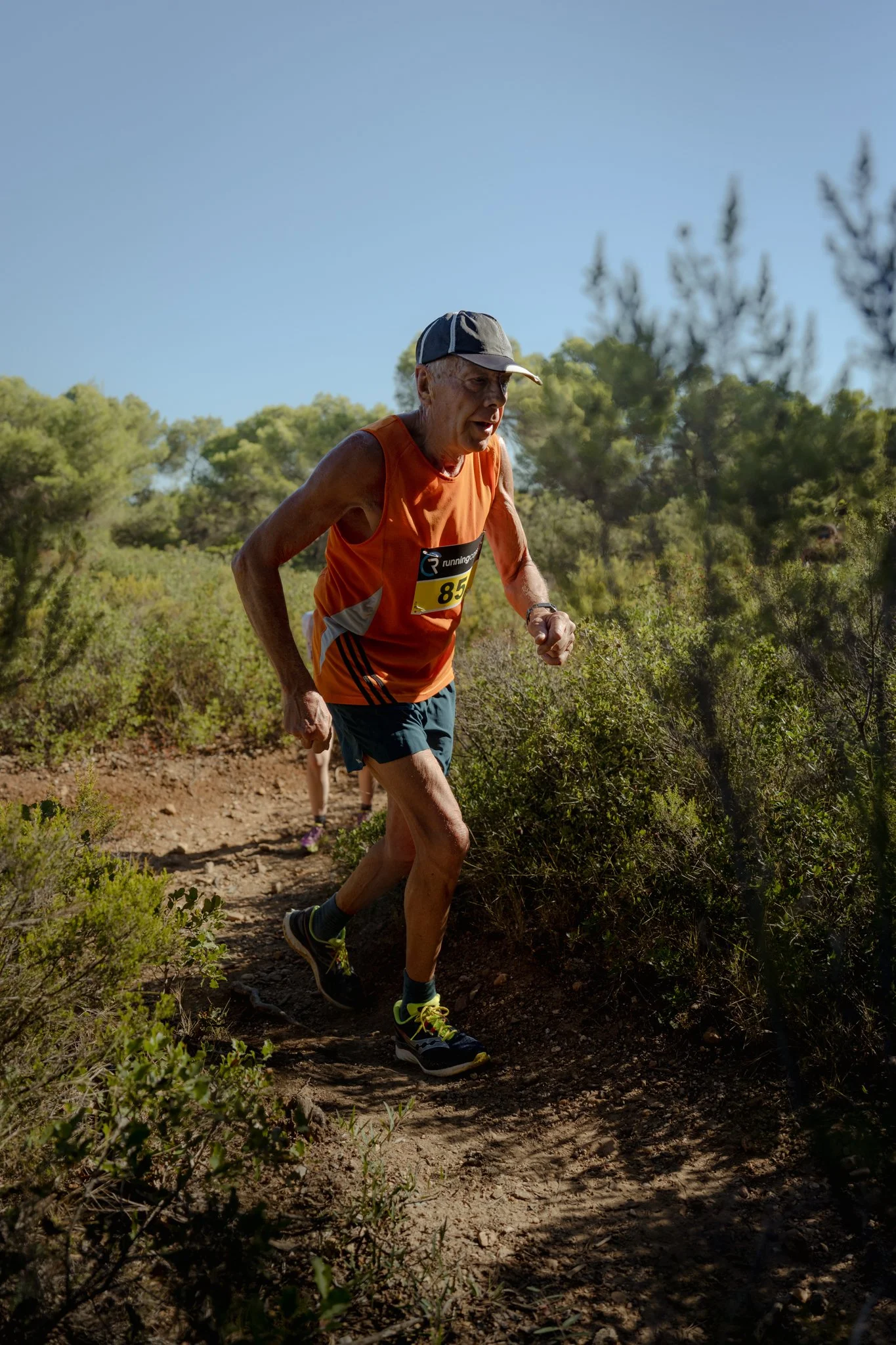 Un homme âgé court sur un sentier en pleine nature sous un ciel bleu, portant un maillot orange, un bonnet et des chaussures de course.