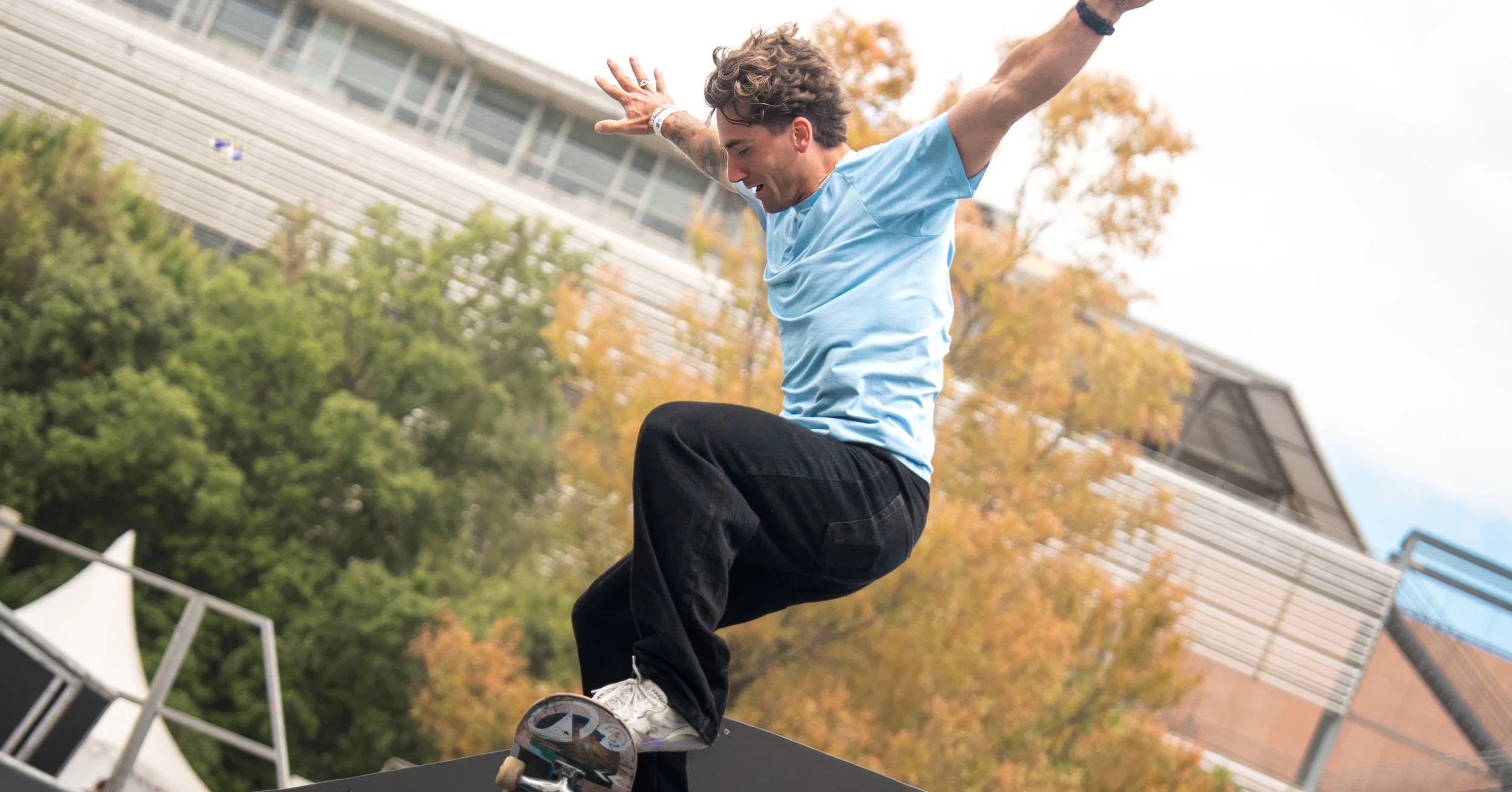 A young man skateboarding outdoors on a black ramp, wearing a light blue t-shirt, black pants, and white sneakers, with trees and a modern building in the background.
