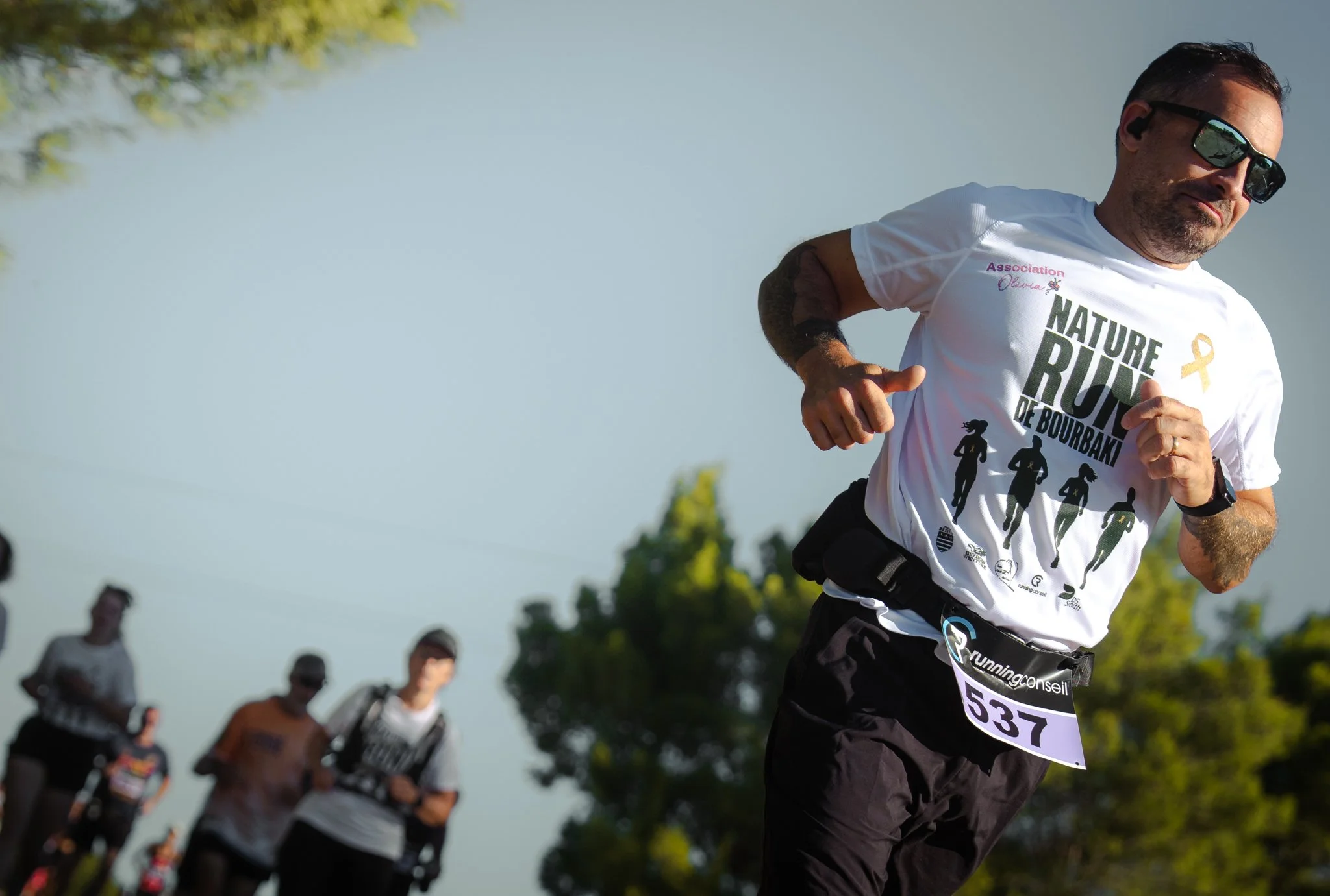 Un homme participant à une course, portant un t-shirt blanc avec le texte "Nature Runners de Bourdais" et une petite broderie jaune. Il court parmi un groupe de coureurs, au grand air avec un ciel clair en arrière-plan.