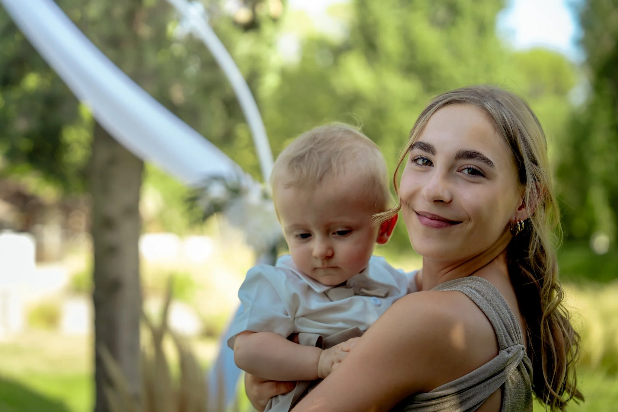 Une jeune femme souriante tient un bébé dans ses bras dans un parc verdoyant avec des arbres en arrière-plan.