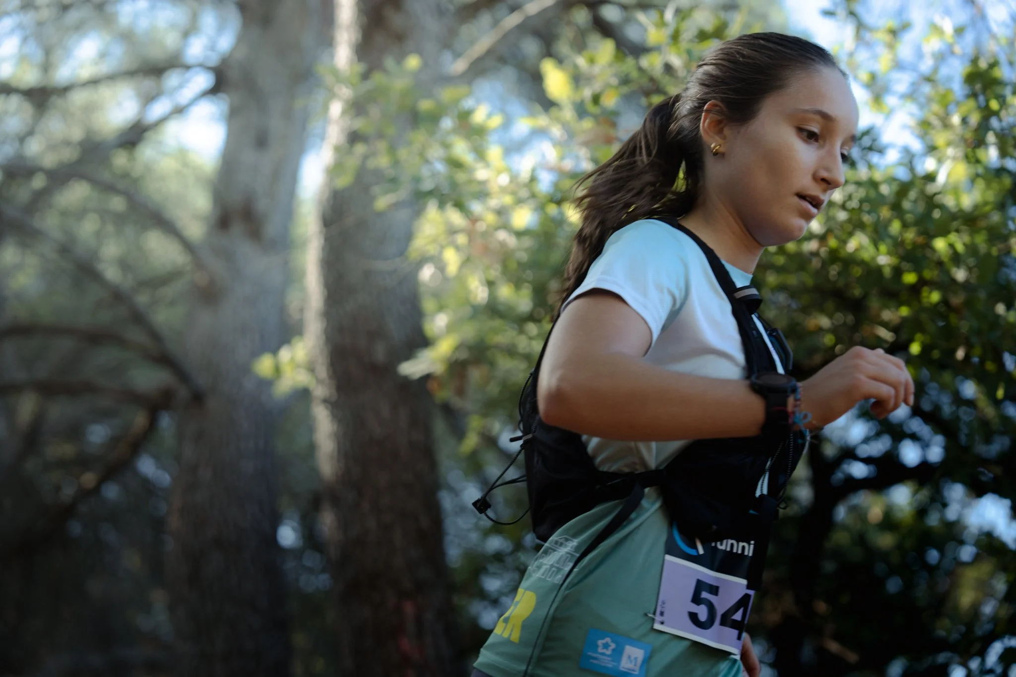 Femme courant dans la forêt lors d'une course ou d'une compétition, portant un gilet d'événement et un numéro de course, avec des arbres en arrière-plan.