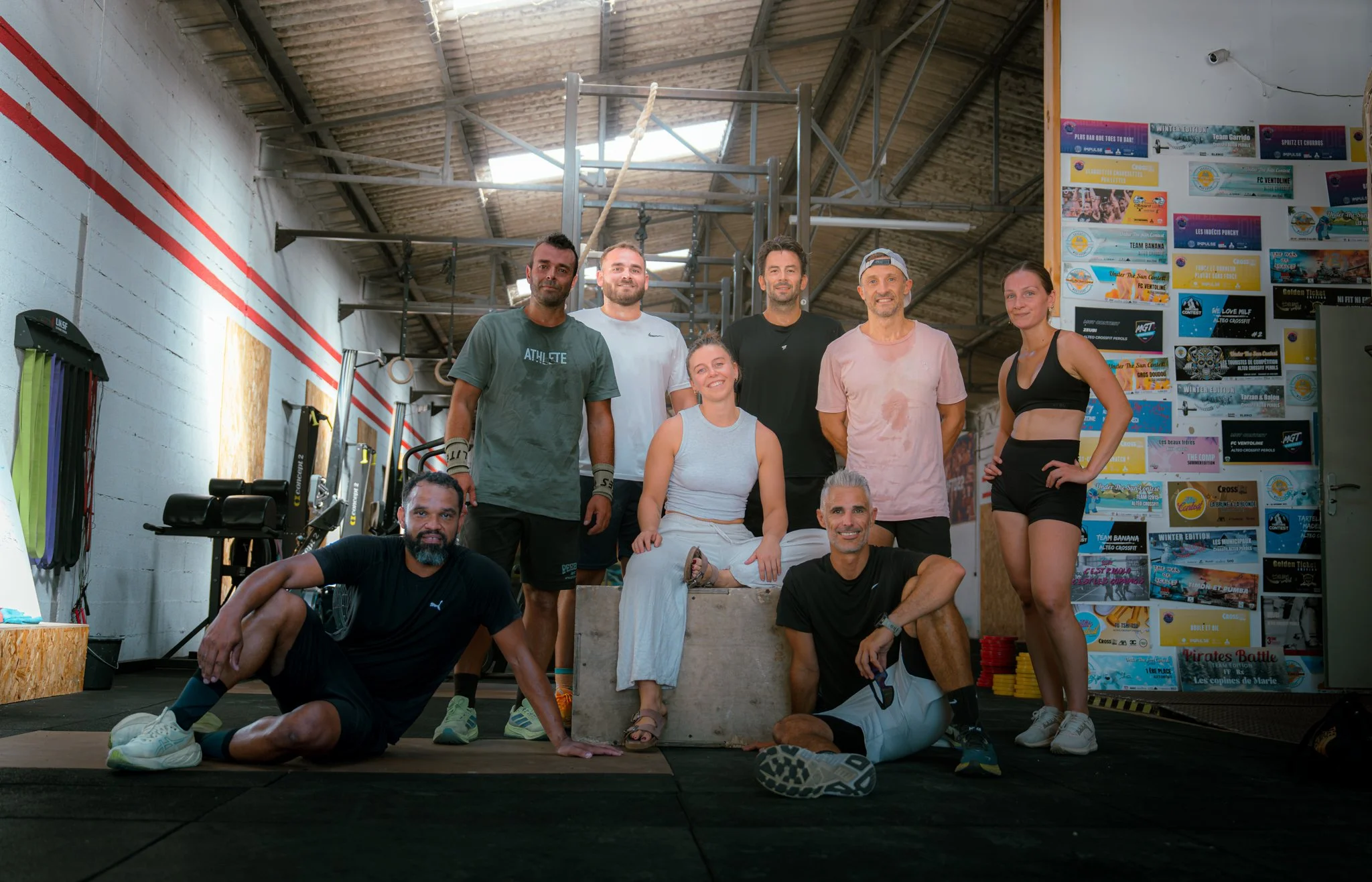 Groupe de personnes dans une salle d'entraînement de sport, posant pour une photo de groupe, certains debout, d'autres assis ou à genoux, avec du matériel de fitness en arrière-plan.