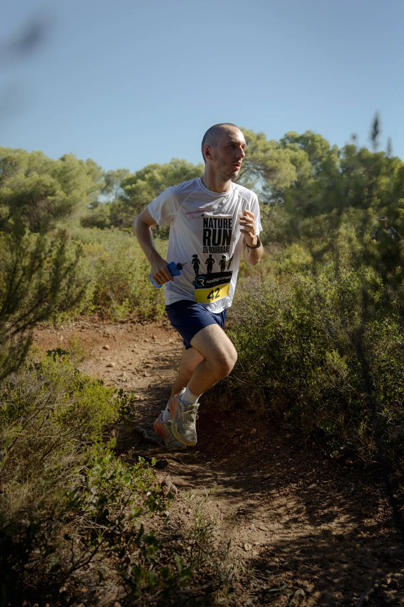Un homme court dans un sentier naturel entouré d'arbustes et d'arbres, portant un t-shirt blanc avec l'inscription "Nature Run" et un numéro de course 42, lors d'une course en plein air par temps ensoleillé.