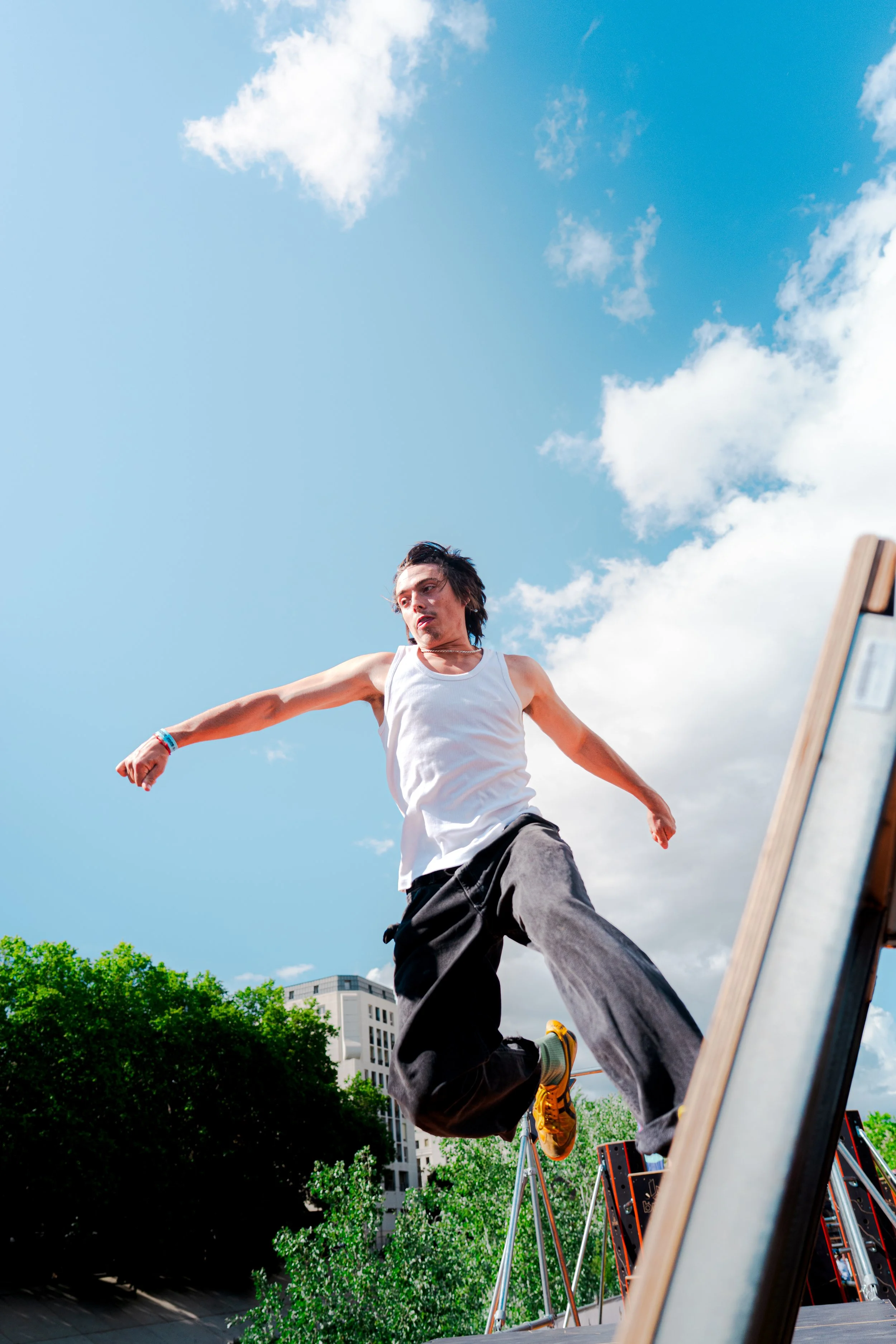 Une personne en train de faire du parkour ou de la mise à l'épreuve de ses compétences en sautant d'une plateforme en plein air, avec un ciel bleu et quelques nuages en arrière-plan et des bâtiments modernes visibles en bas, sous un arbre vert dense.