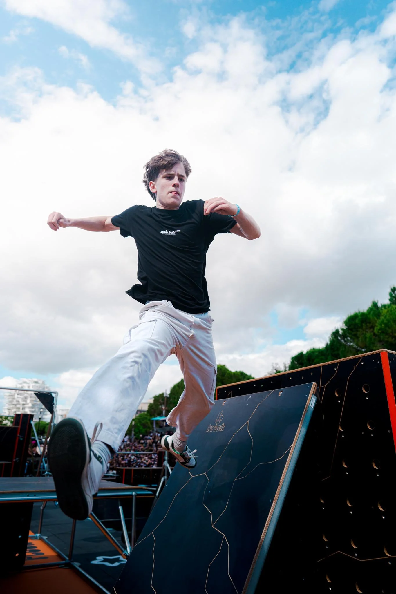 A man in a black t-shirt and white pants falls backward off an obstacle during a competitive event, with a crowd watching and a blue sky with clouds overhead.