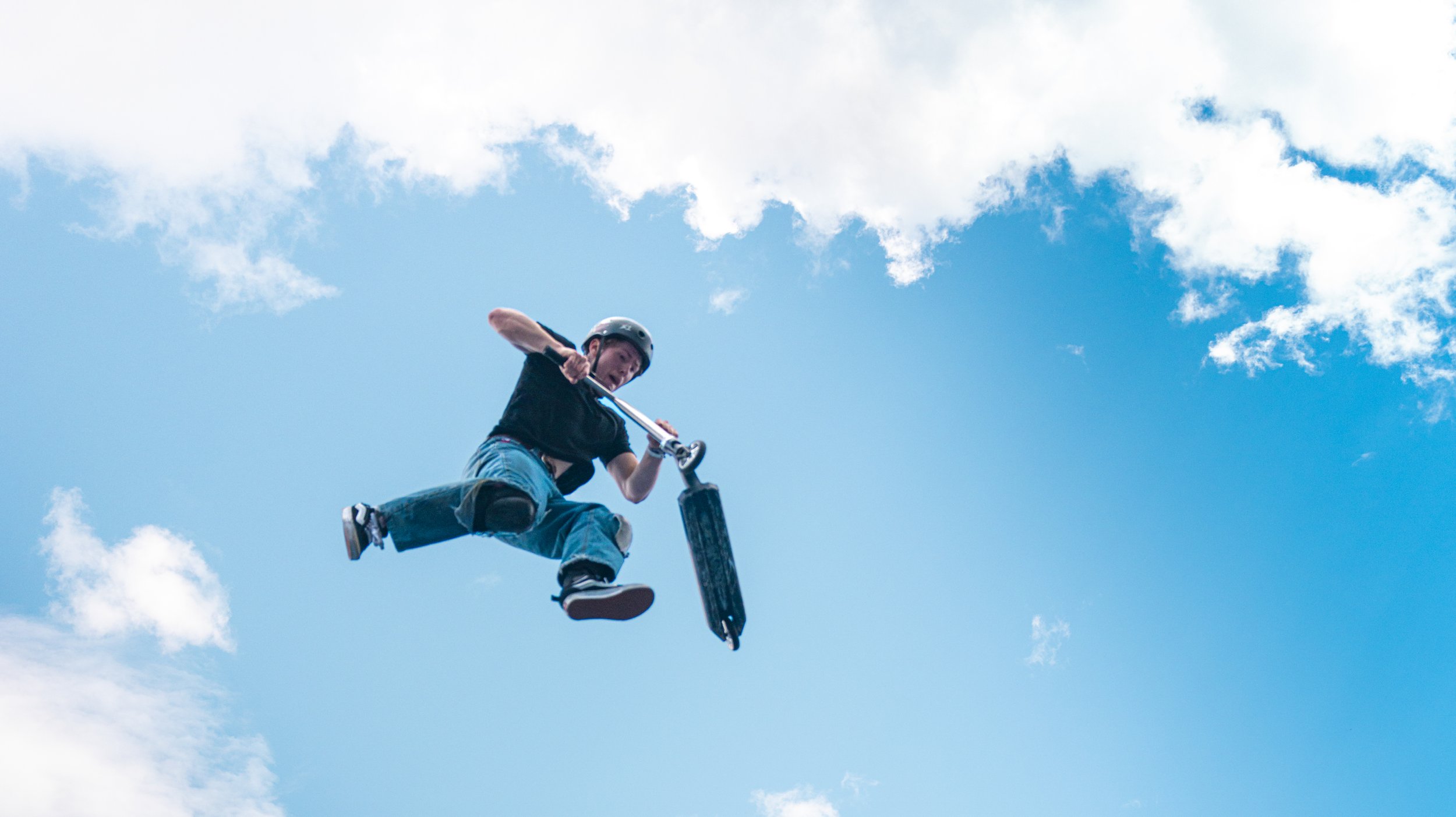 Person riding a scooter mid-air against a blue sky with clouds.