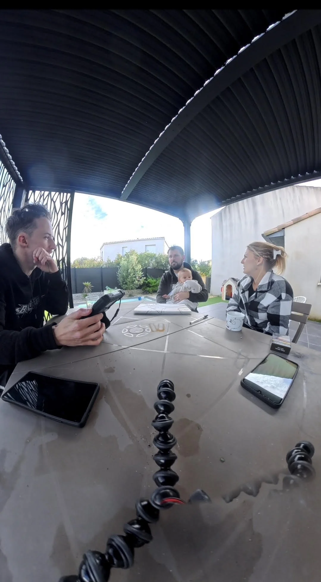 Groupe de quatre personnes, deux hommes, une femme et un bébé, assis autour d'une table sous une pergola, avec divers appareils électroniques et une caméra sur un trépied, en extérieur avec un jardin en arrière-plan.