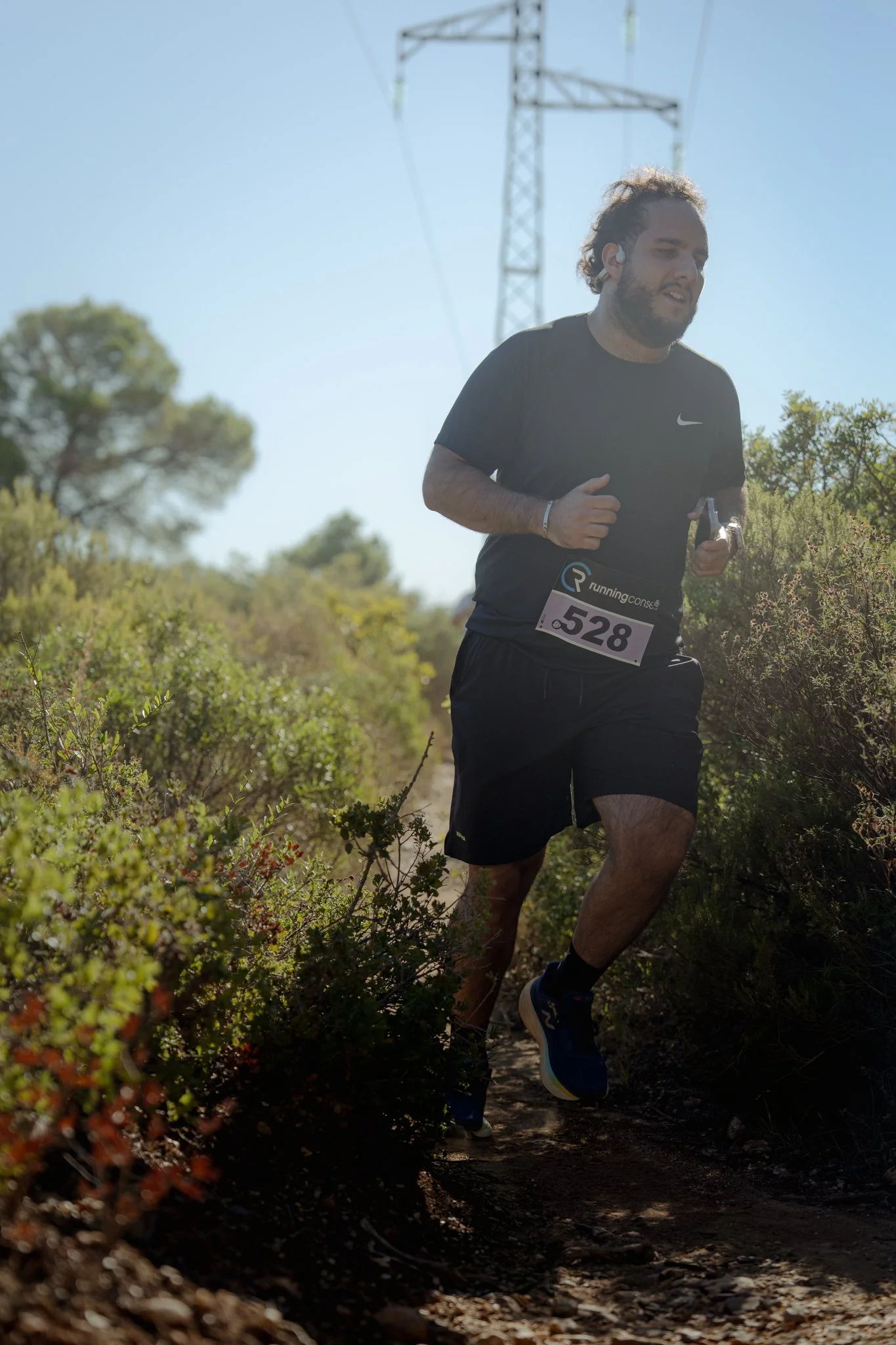 Un homme court en pleine nature, portant un t-shirt noir, un numéro de course 528, et écoute des écouteurs. Le soleil brille et il y a des arbustes autour du sentier.