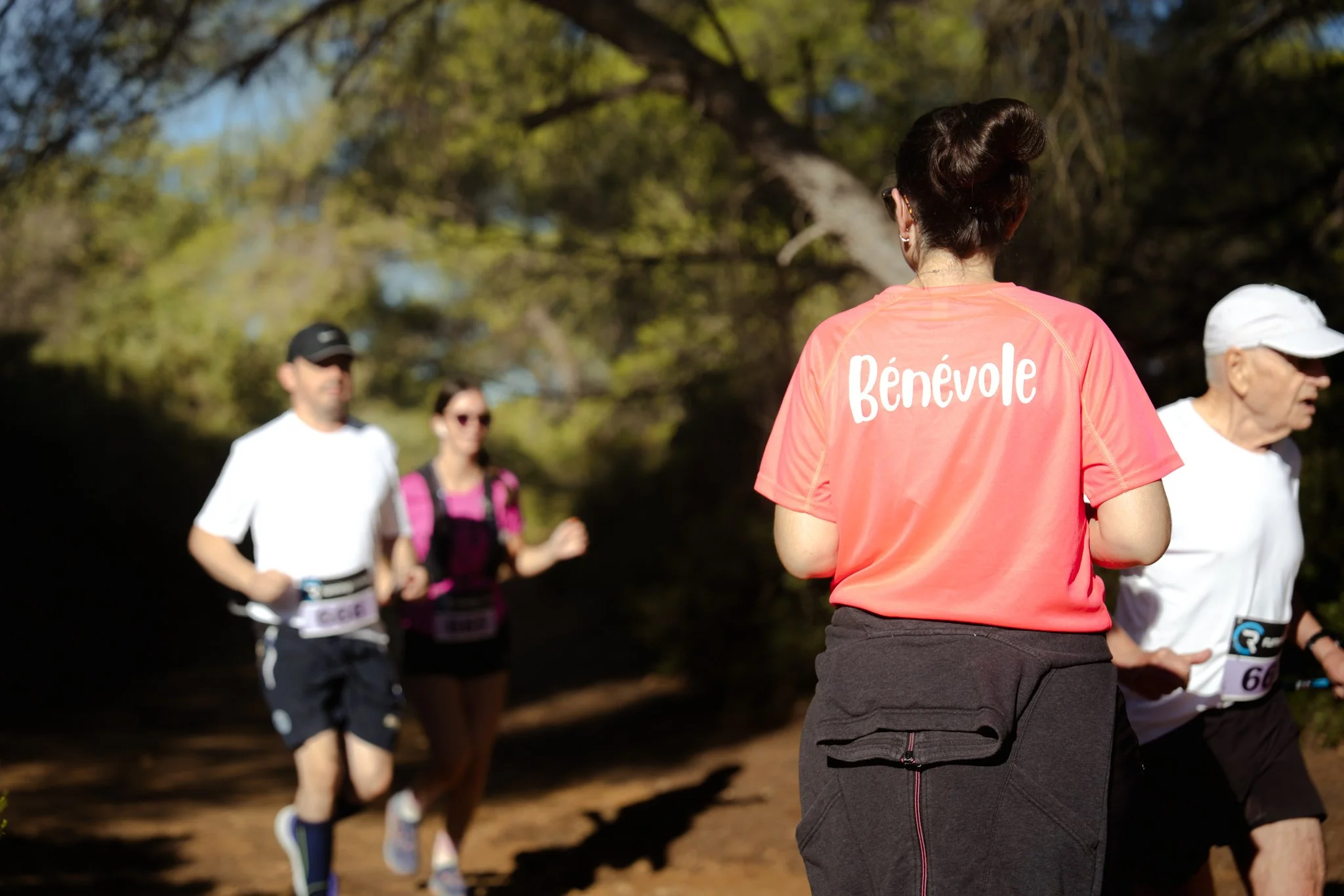 Une bénévole regarde un groupe de coureurs lors d'une course en extérieur dans un parc, avec des arbres en arrière-plan, en journée ensoleillée.
