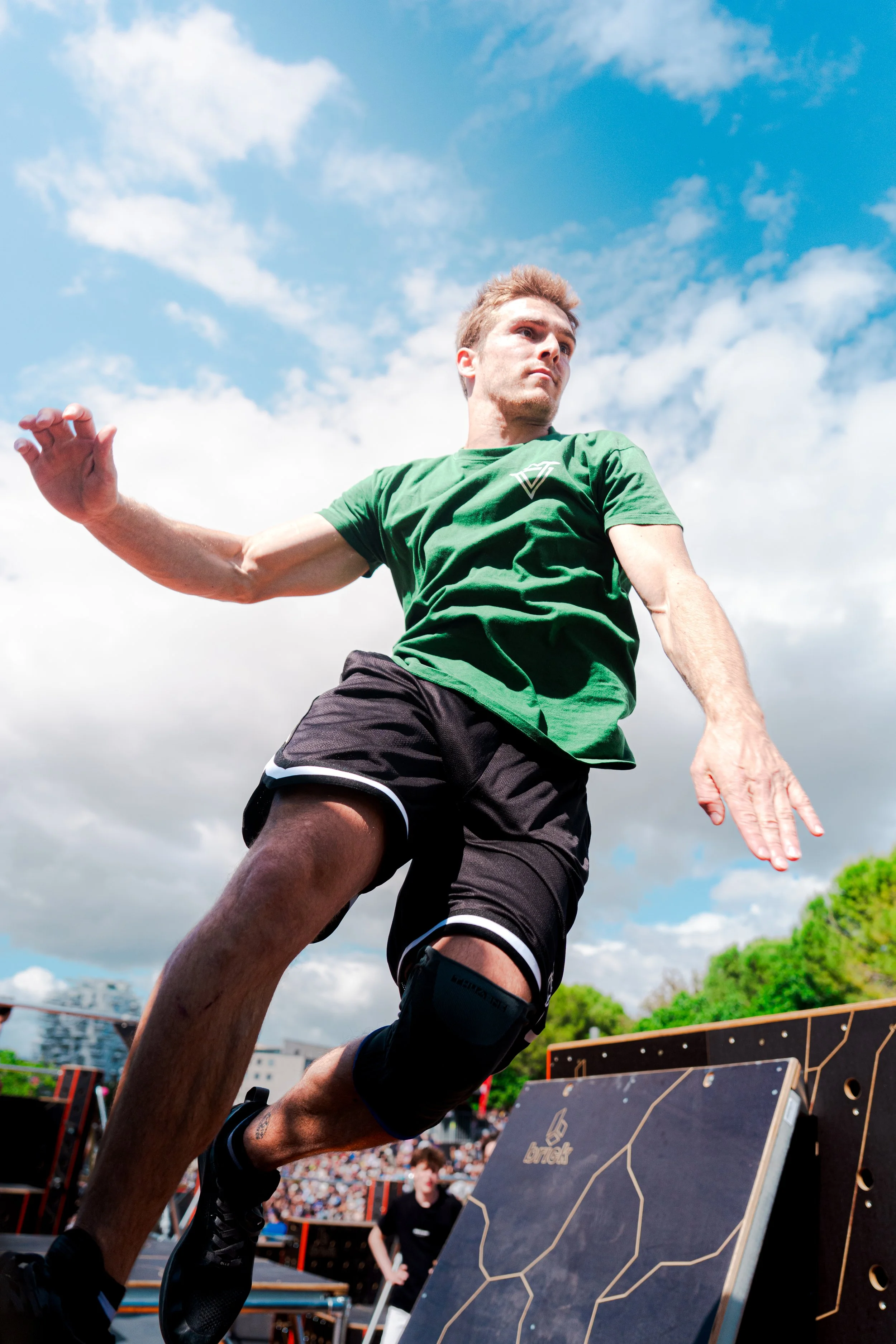 A male athlete in a green shirt and black shorts, wearing a knee brace, is mid-air during an obstacle course competition outdoors under a partly cloudy sky, with spectators and obstacle equipment visible in the background.