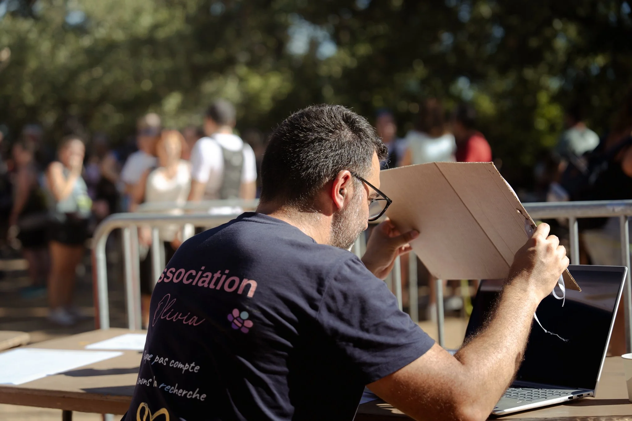 Un homme avec des lunettes et une barbe, portant un t-shirt noir avec des écritures roses, assis à une table en extérieur, tenant une planche en carton devant son visage et travaillant sur un ordinateur portable. En arrière-plan, plusieurs personnes 