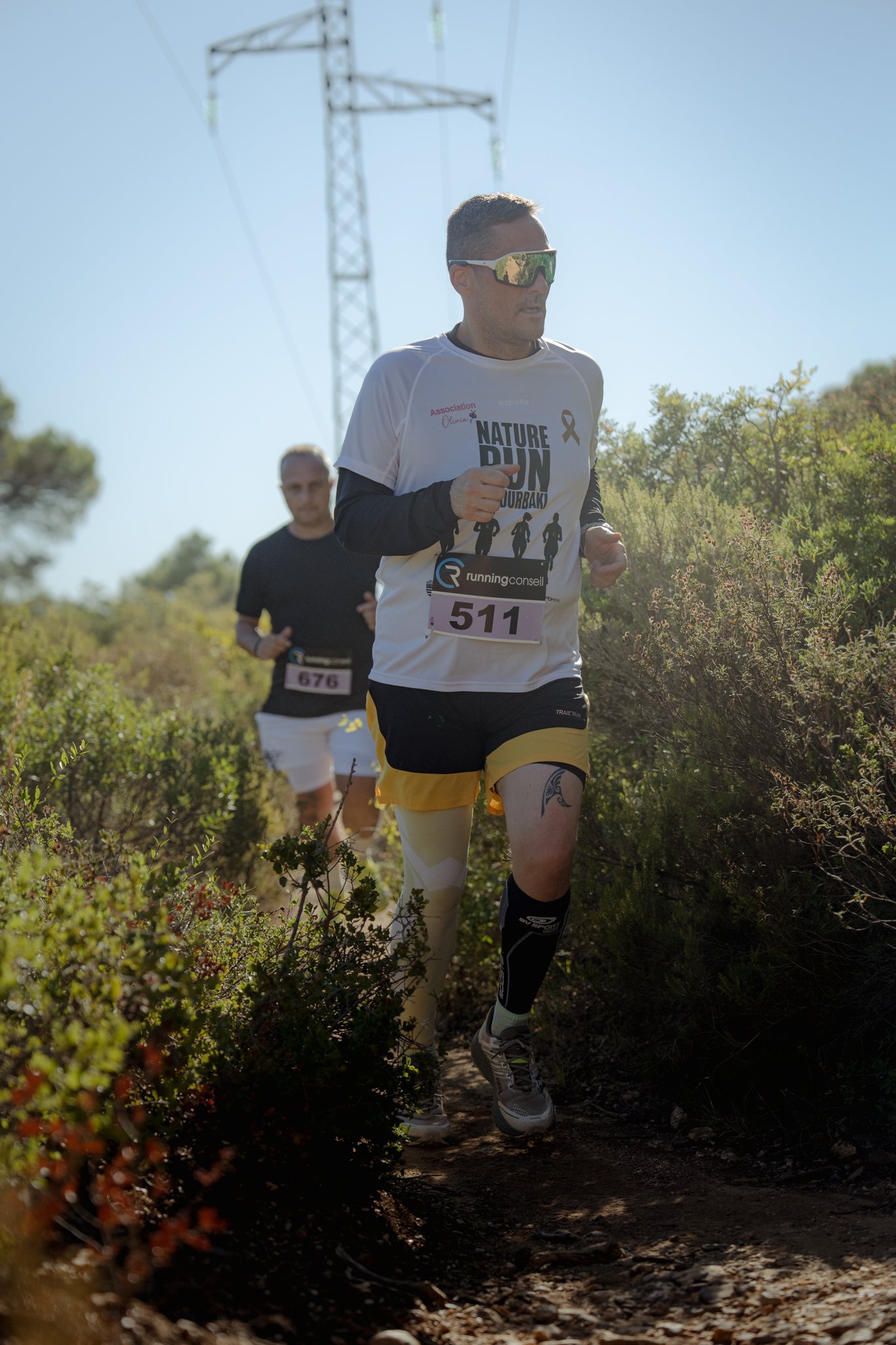 Deux coureurs participent à une course en forêt, portant des maillots avec des numéros, chemins de terre et végétation dense.