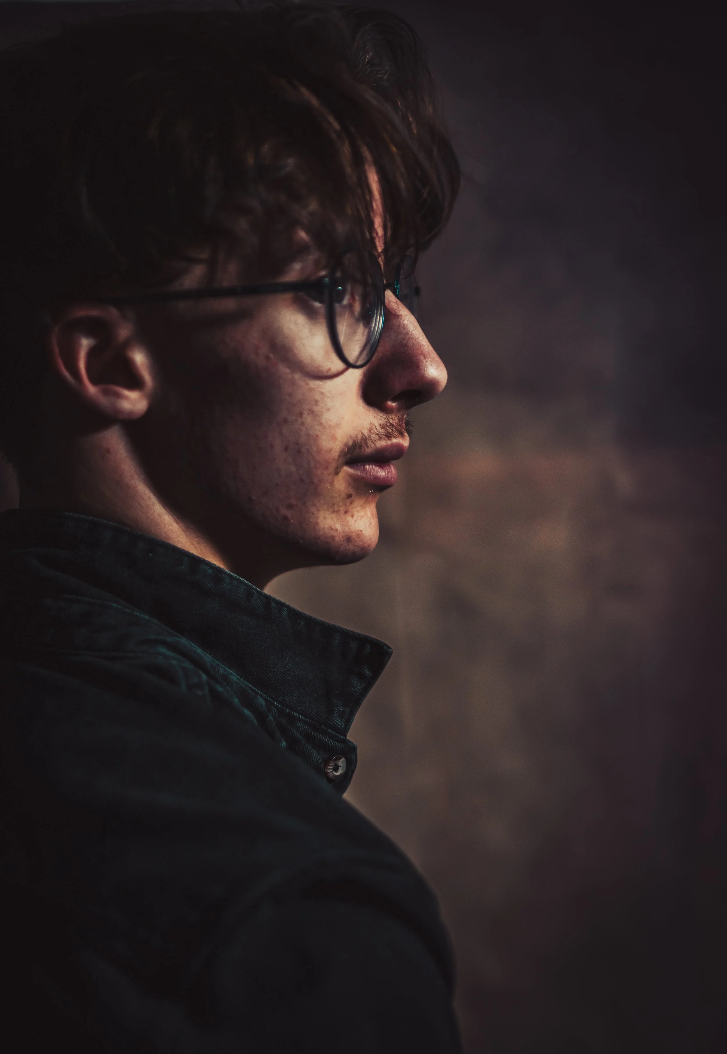 Portrait d'un homme âgé regardant vers la droite, portant des lunettes et une veste noire, dans un environnement sombre.