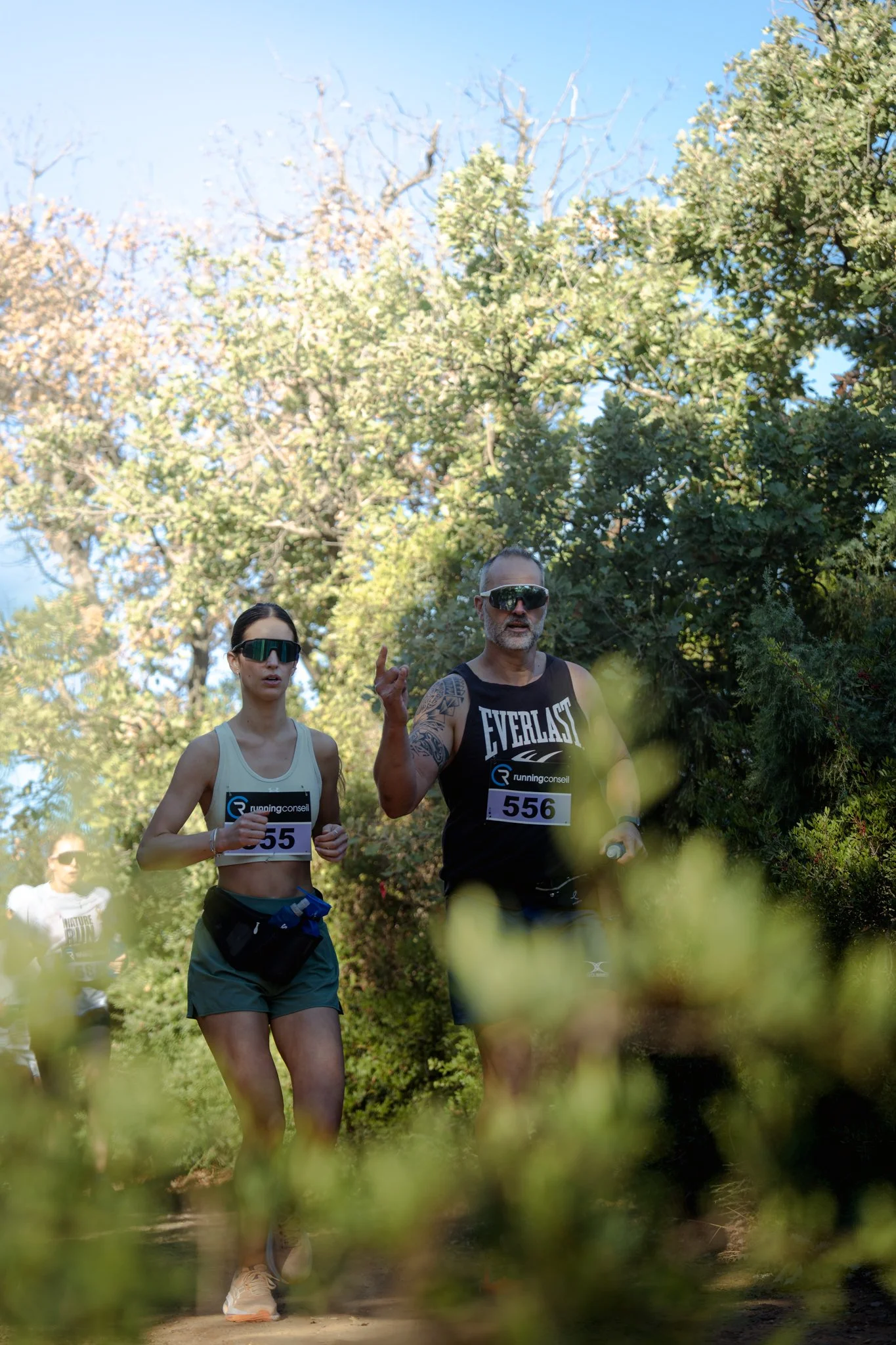 Deux personnes courent dans un parc en tenue de sport, portant des dossards de course, entourées d'arbres avec un ciel bleu en arrière-plan.