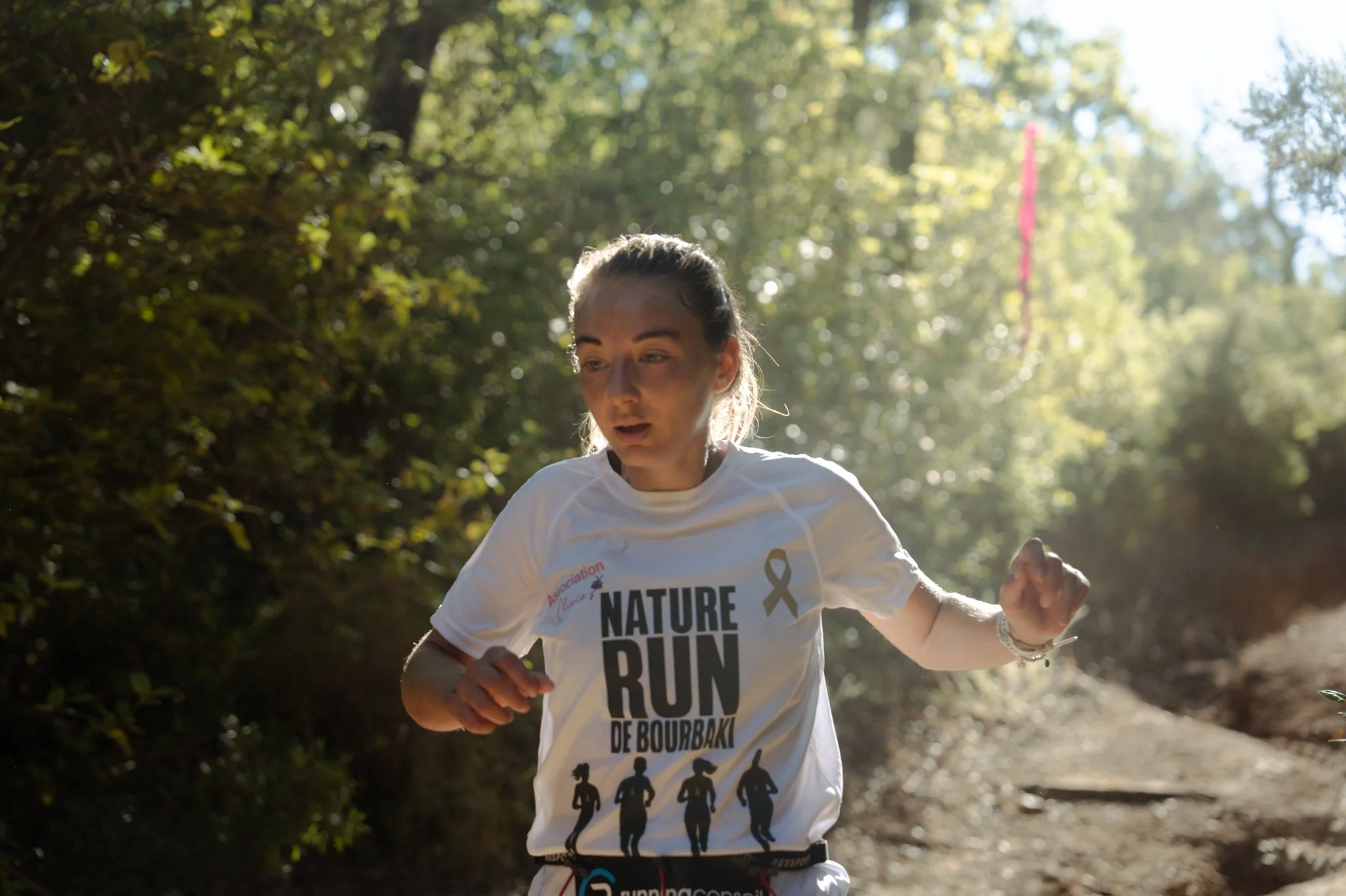 Une femme participe à une course à l'extérieur, portant un t-shirt blanc avec le texte 'NATURE, RUN, DE BOURBAKI'. Elle court dans une voie bordée d'arbres, avec de la lumière solaire filtrant à travers le feuillage.