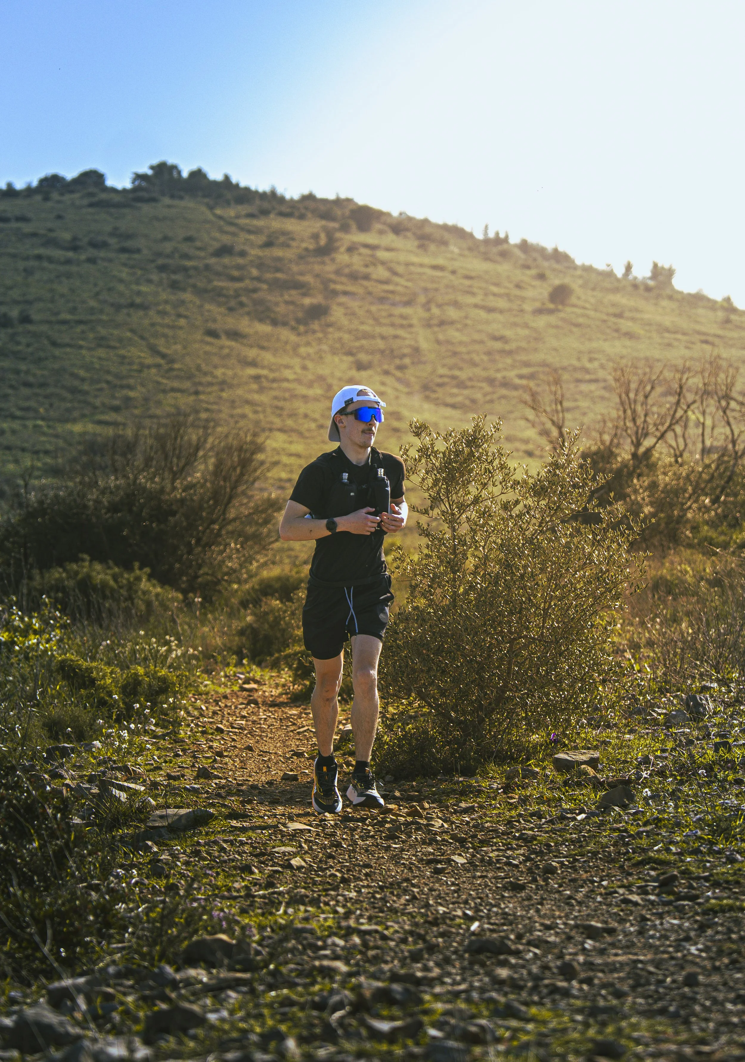 Homme en tenue de course à pied, portant un chapeau blanc, des lunettes de soleil bleues, une montre et une bouteille d'eau, courant sur un sentier de randonnée dans un environnement nature avec des collines et des arbustes, au coucher ou lever du soleil